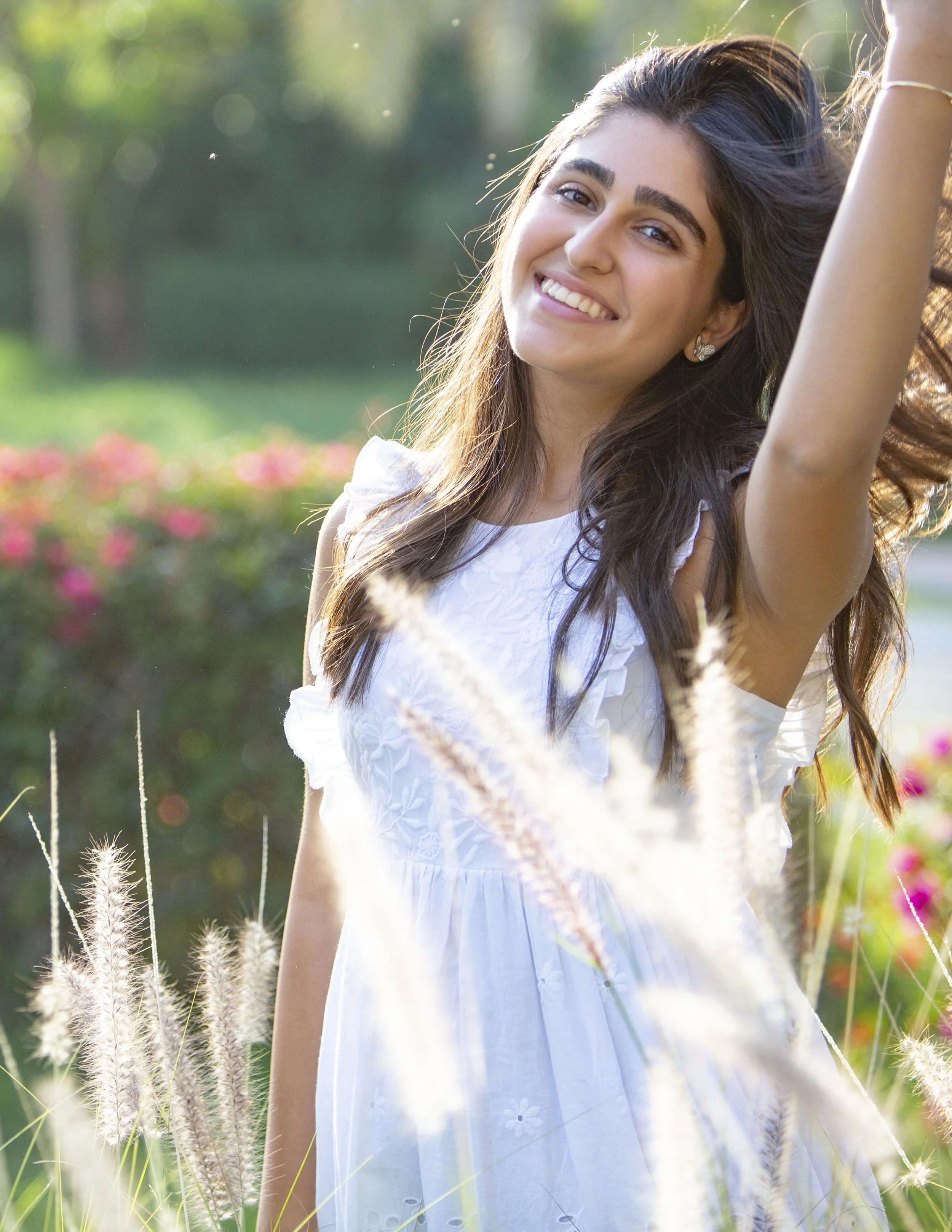 Smiling young woman in a white dress standing outdoors in a garden during daytime with sunlight, surrounded by tall grasses and flowers.