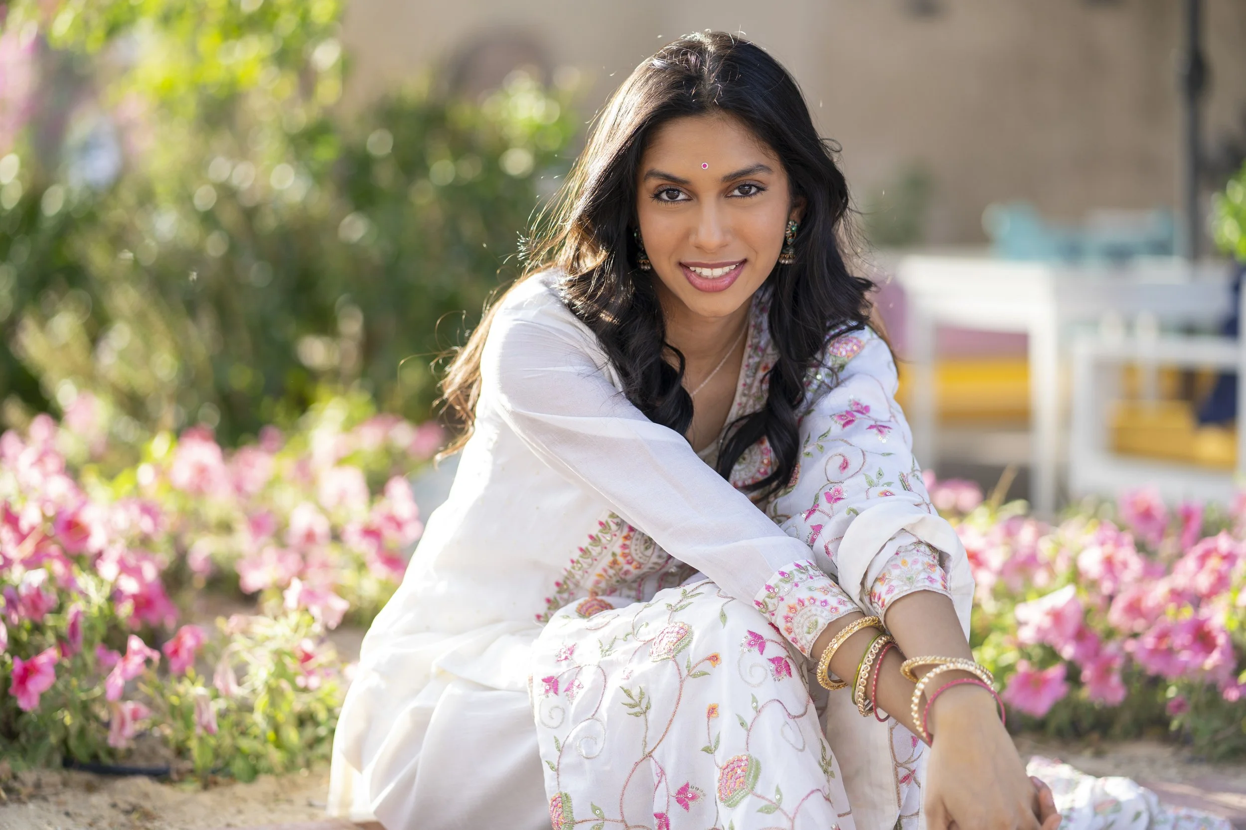 A woman with long black hair, dressed in a white traditional outfit with floral embroidery, sits outdoors among pink flowers, smiling at the camera.