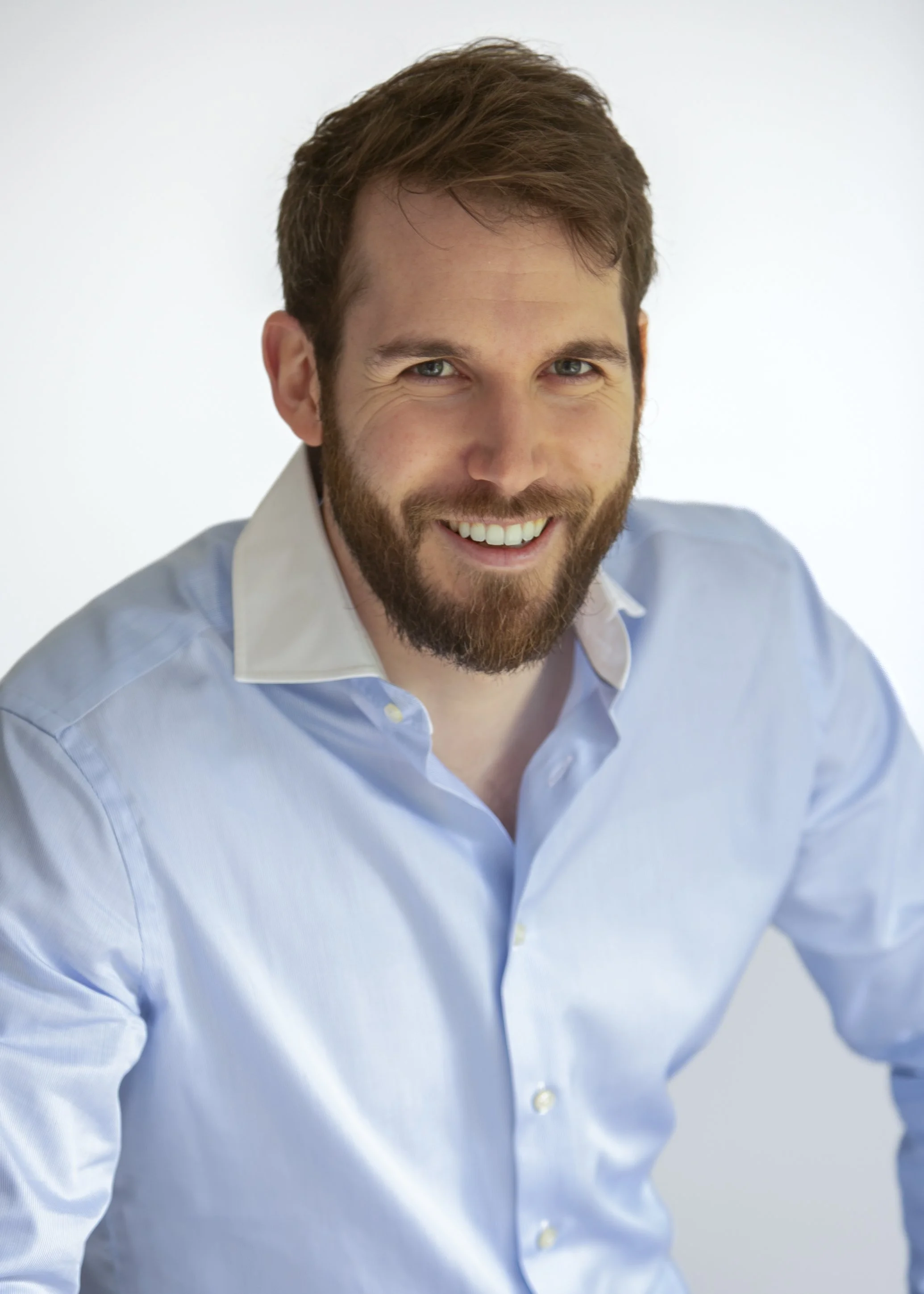 A smiling man with brown hair and a beard, wearing a light blue dress shirt with a white collar, in front of a plain white background.