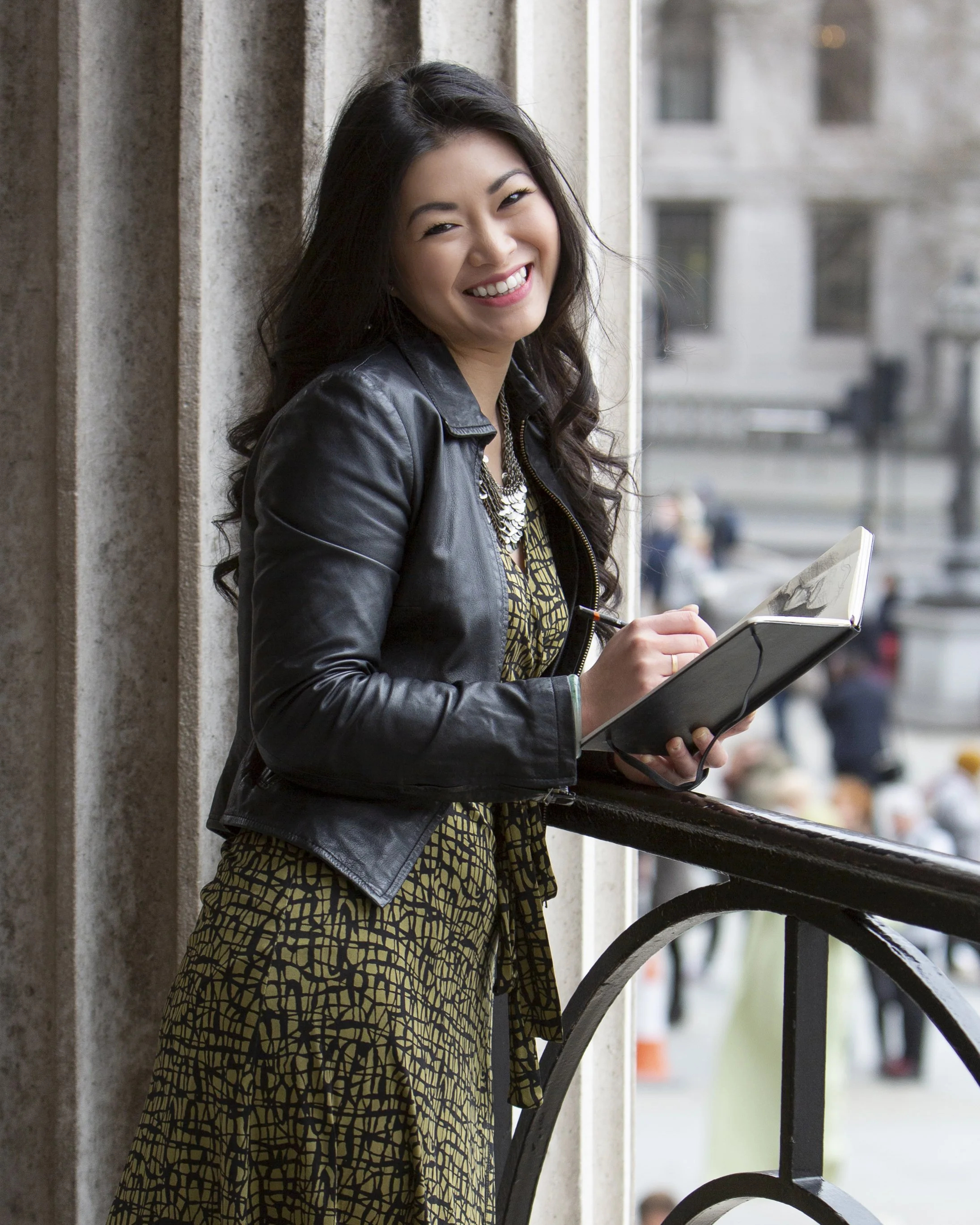 A young woman with long dark hair and a bright smile, wearing a black leather jacket, a patterned dress, and holding a notebook, standing on a balcony in an urban setting.
