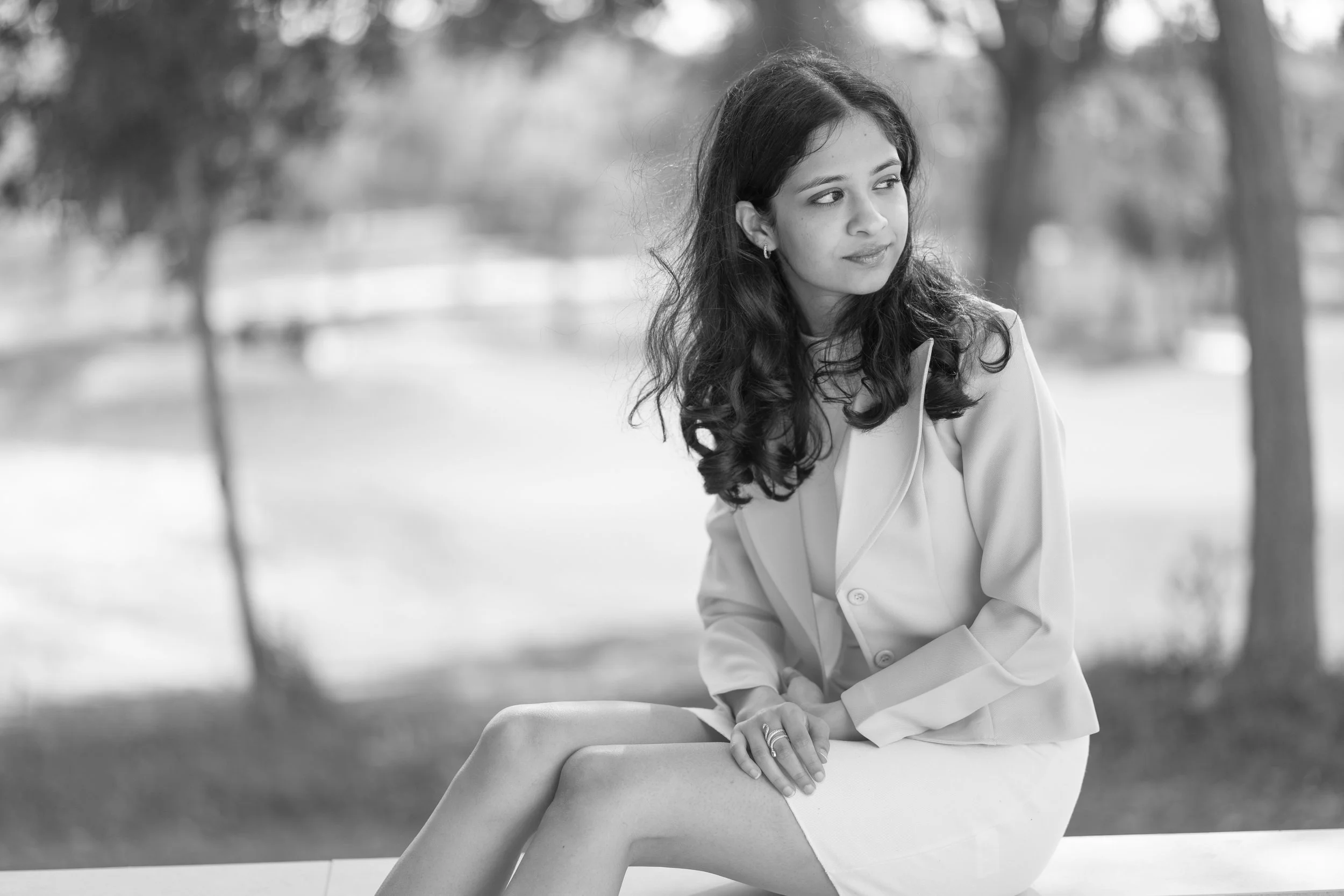 A woman with dark, curly hair sitting outdoors on a bench in a park, wearing a light-colored jacket and skirt, with trees and a blurred background.
