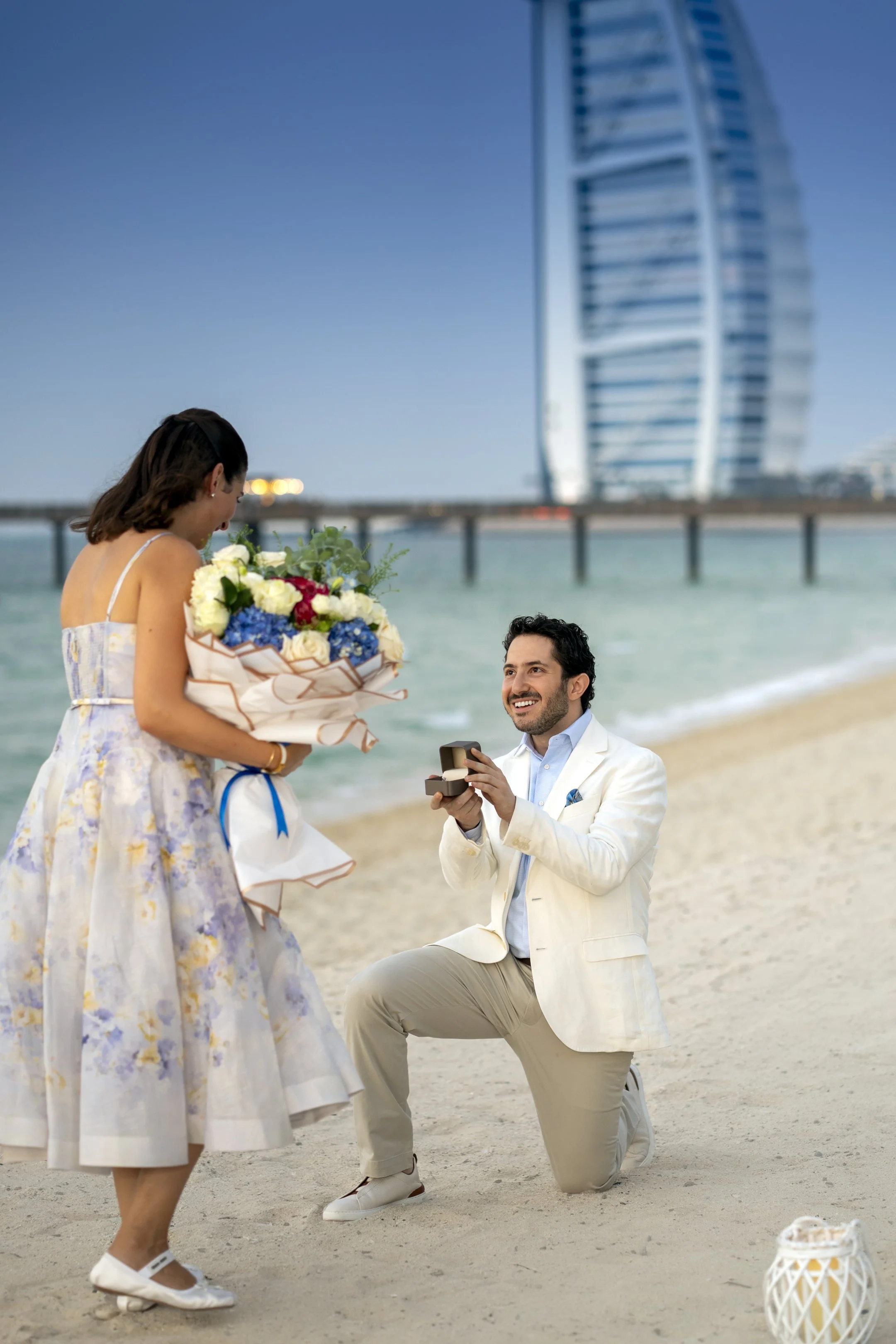 A man proposing marriage to a woman on a beach, with the man kneeling and holding a ring box, the woman holding a large bouquet of flowers, and a modern skyscraper in the background.