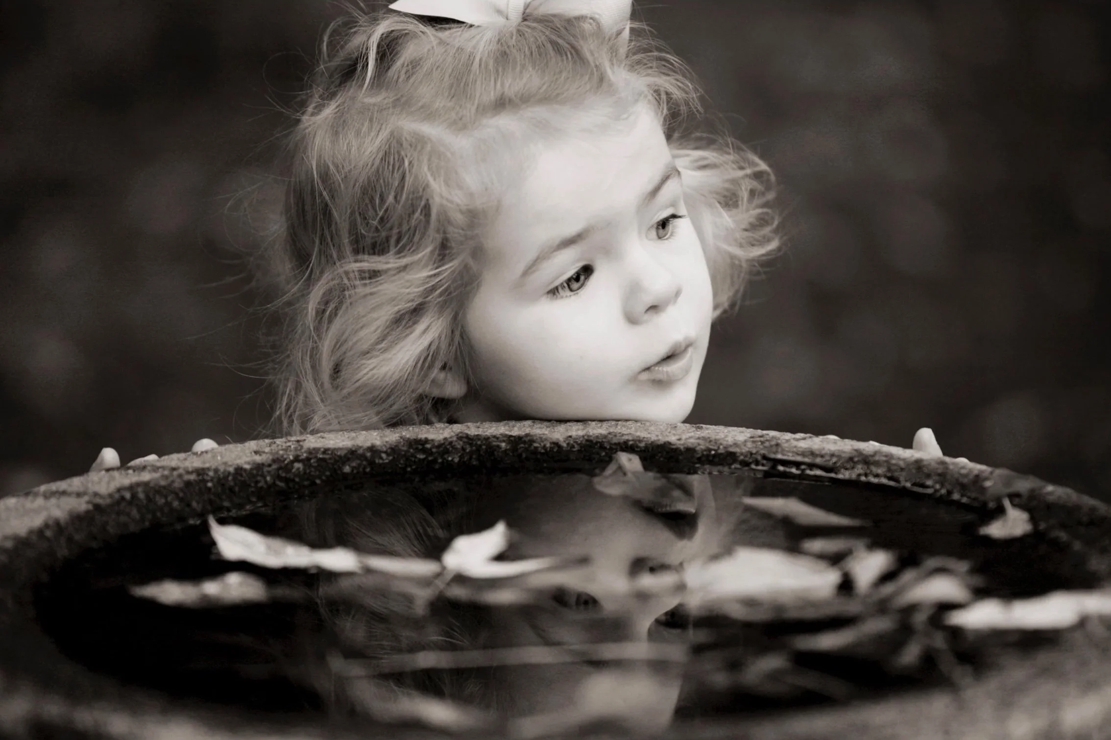 Close-up of a young girl with curly hair and a bow in her hair, looking at a pond with floating leaves.