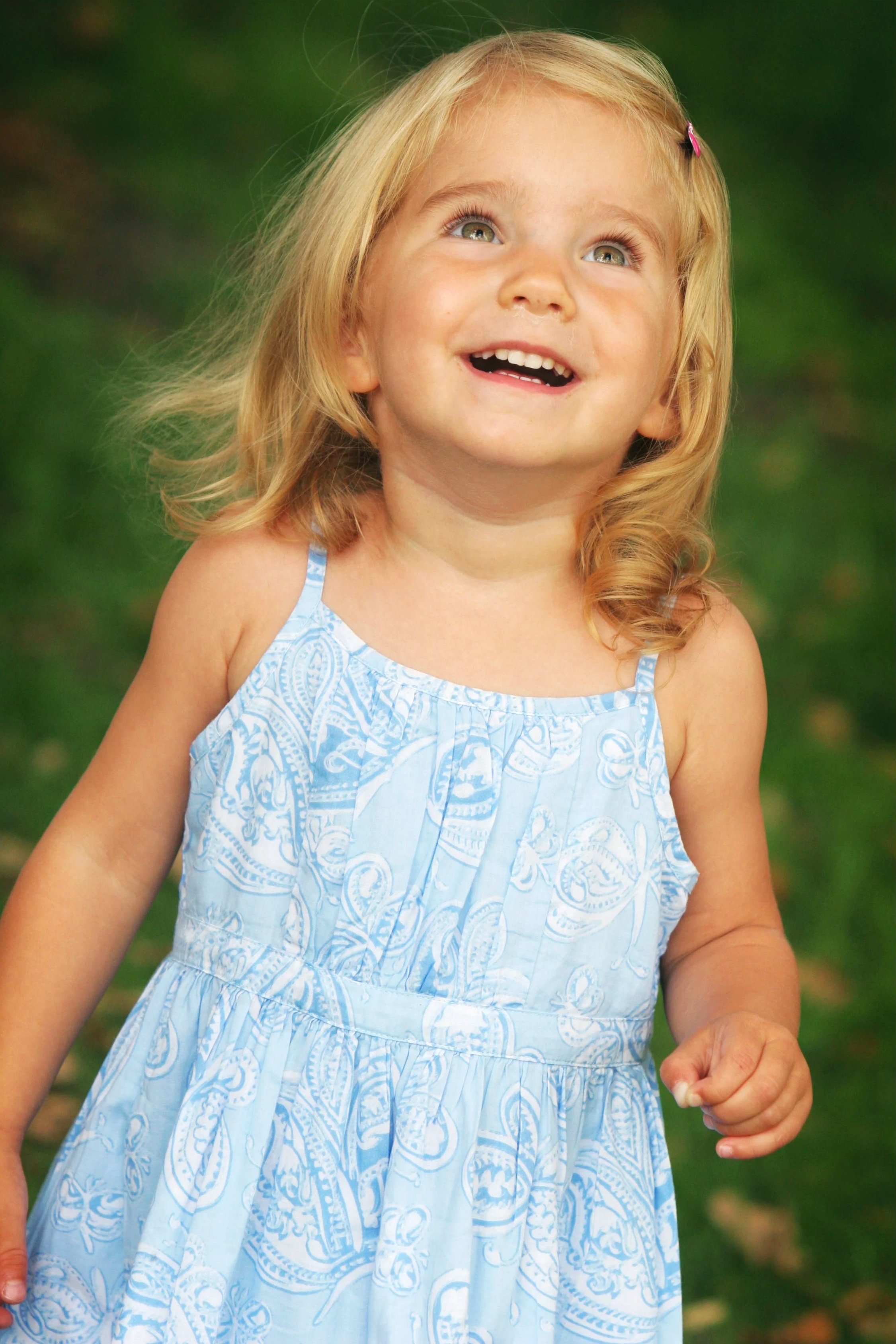 A young girl with blonde hair, wearing a light blue patterned dress, smiling and looking up outdoors with a blurred green background.