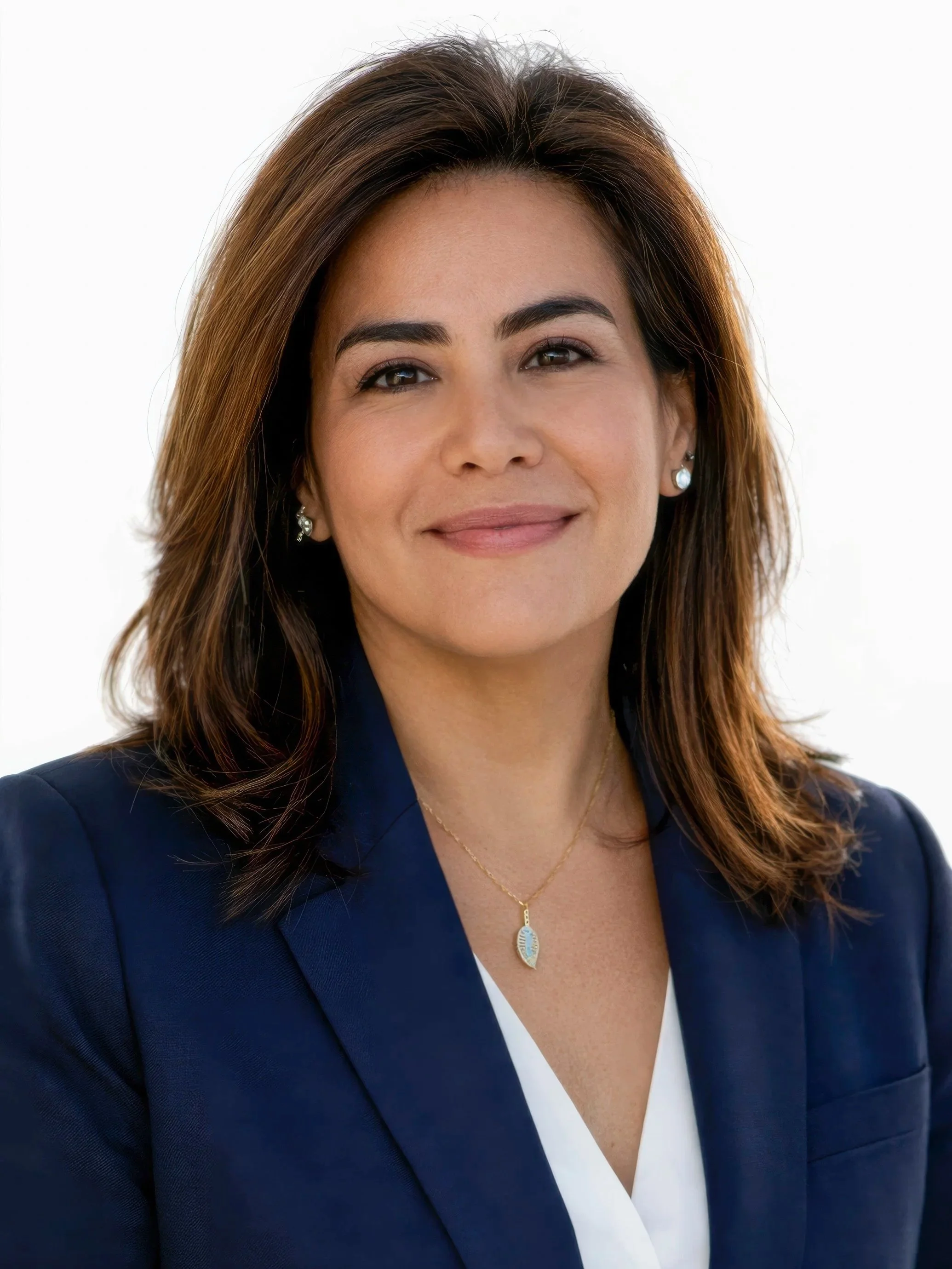 A woman with shoulder-length brown hair, wearing a dark blue blazer, white shirt, silver earrings, and a gold necklace with a blue and white oval pendant, smiling slightly against a plain white background.