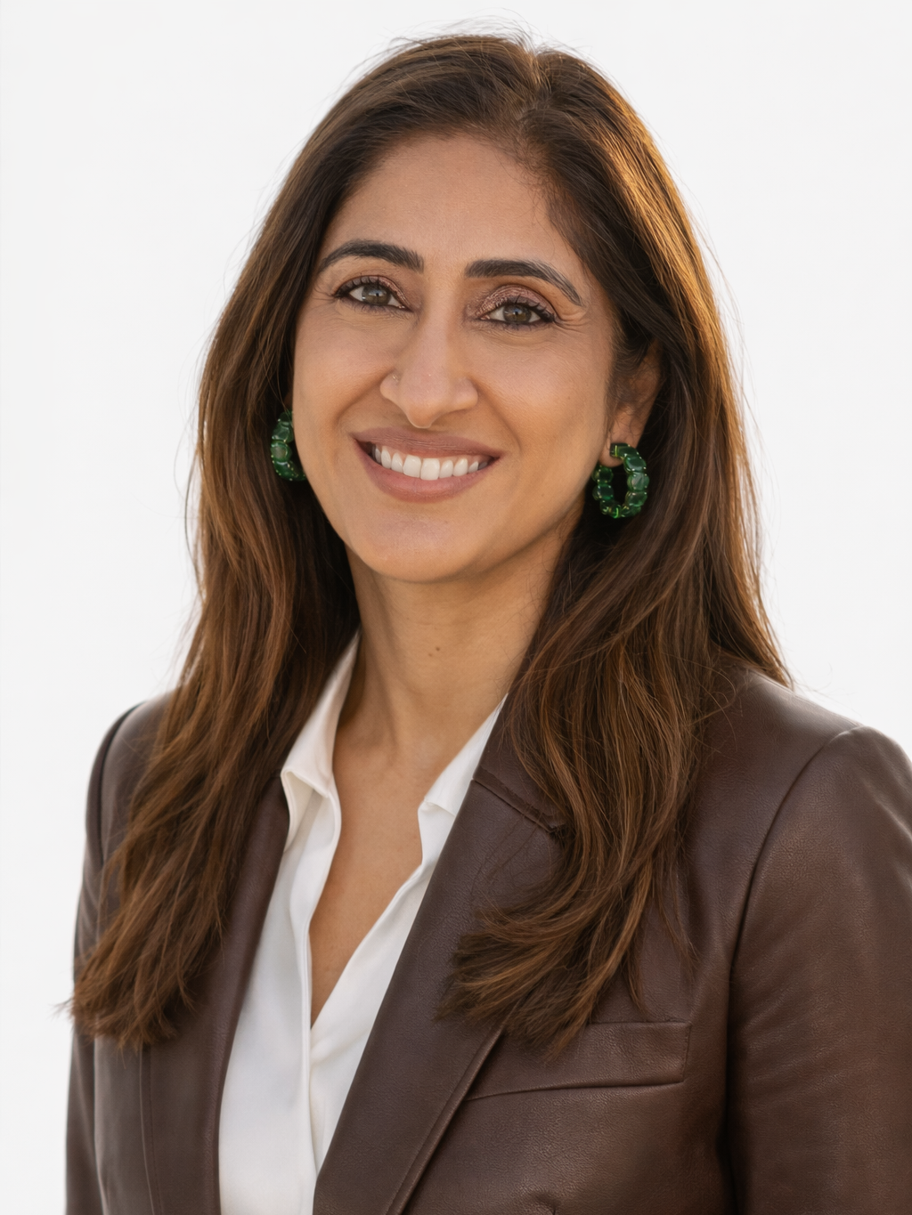 A woman with long brown hair, green earrings, wearing a white shirt and a brown leather jacket, smiling at the camera.