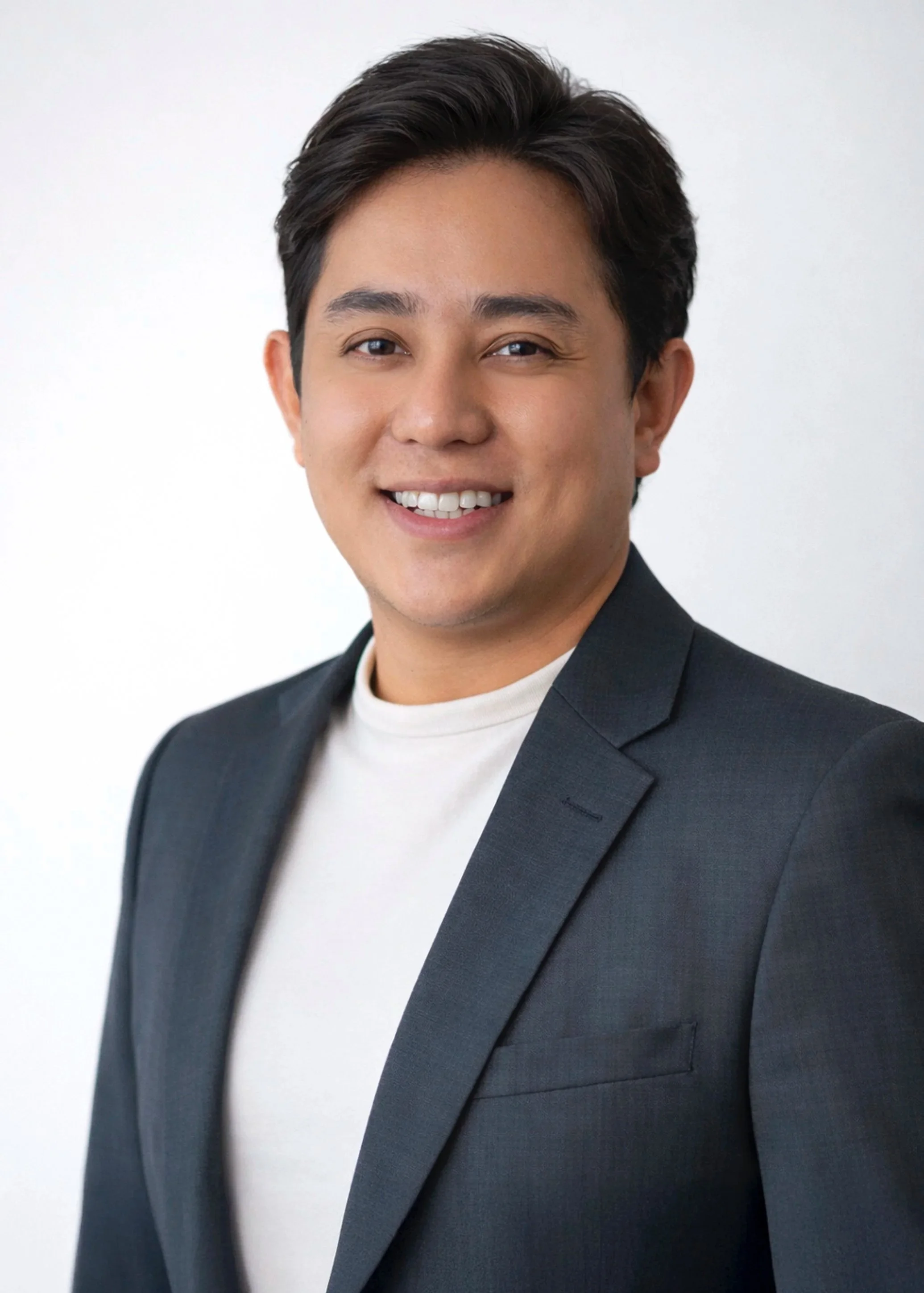 Headshot of a young man with dark hair, wearing a dark blazer and white shirt, smiling against a plain white background.