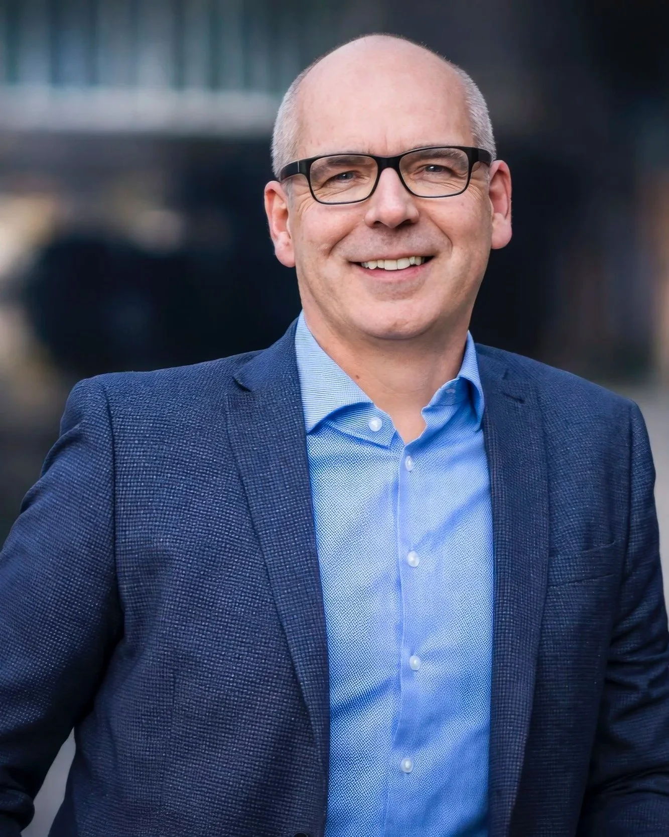 A smiling middle-aged man with glasses, dressed in a blue collared shirt and navy blazer, standing outdoors with a blurred cityscape background.