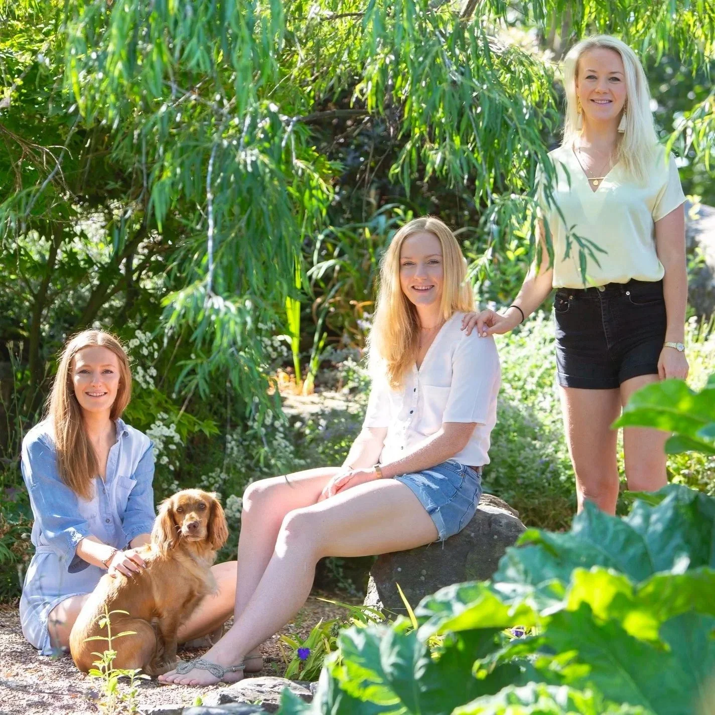 Three women and a dog outdoors in a lush garden or park with green foliage. One woman is sitting on a rock with her legs crossed, another woman is kneeling beside her, and the third woman is standing outside the group. The women are smiling and appea