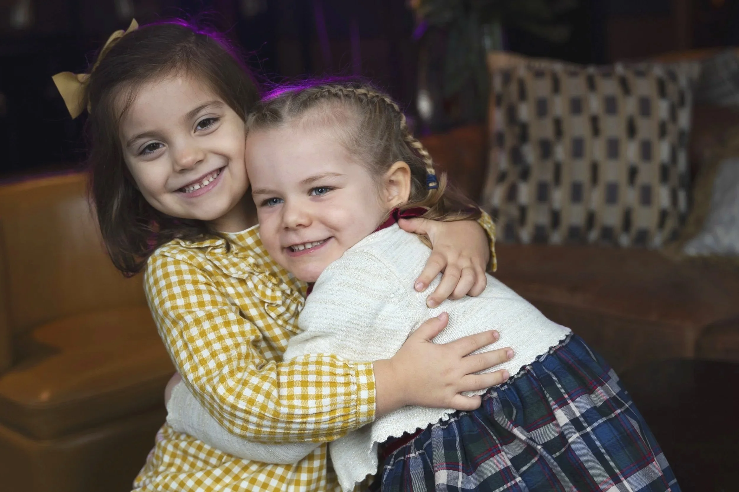 Two young girls hugging each other and smiling indoors, one with dark hair and the other with light hair, both wearing checkered clothing.