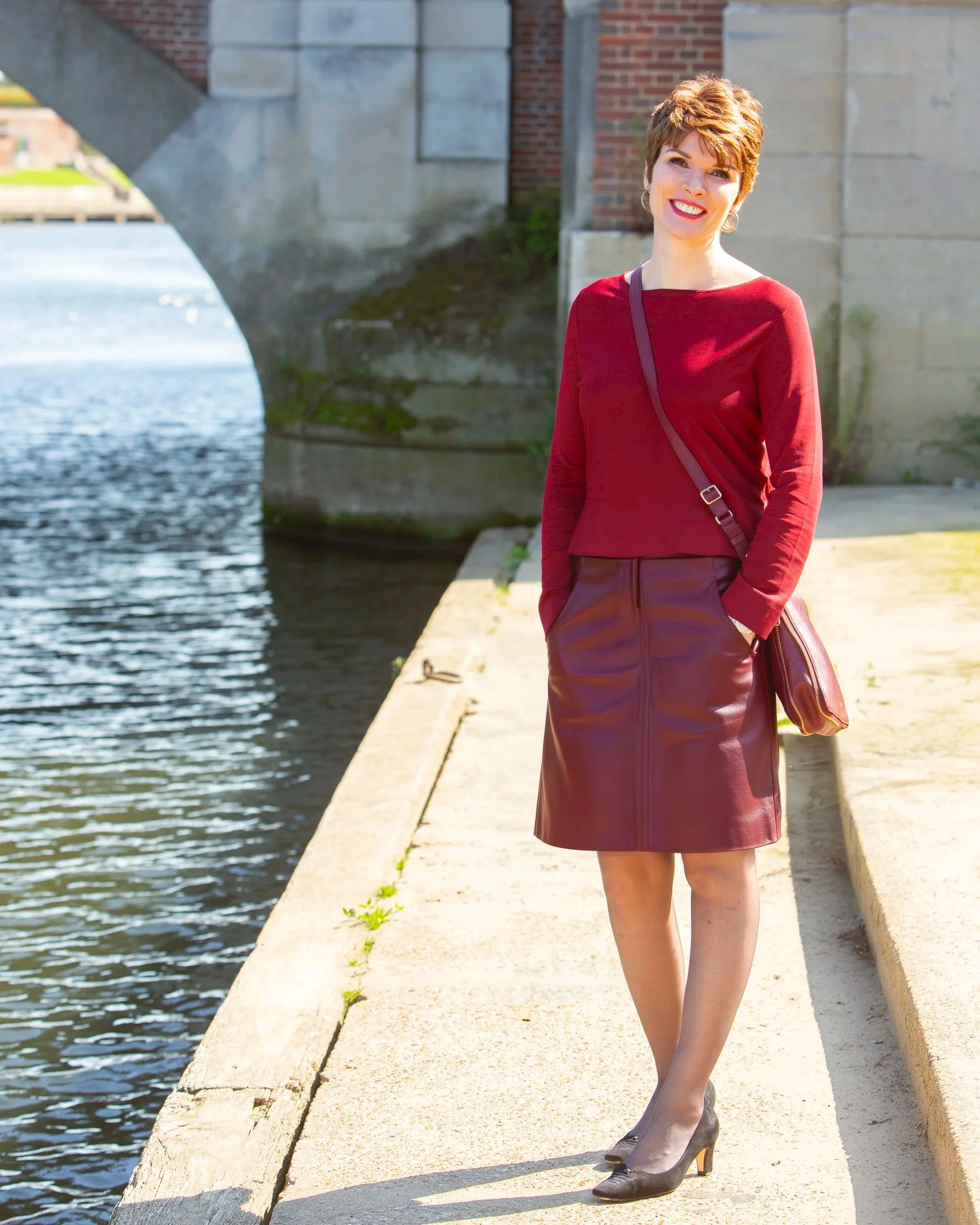 A woman with short auburn hair wearing a red long-sleeve top, a red leather skirt, and black heels stands near a canal under a bridge, smiling at the camera