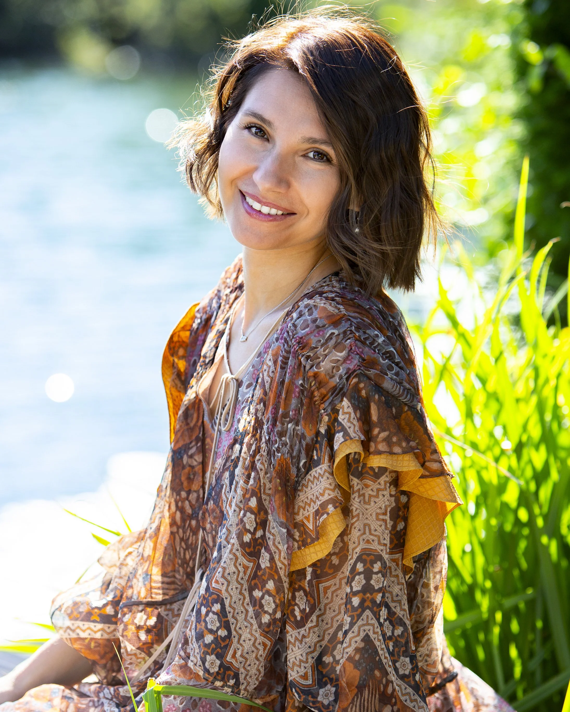 Smiling woman with short, wavy brown hair sitting outdoors near water and green plants on a sunny day.