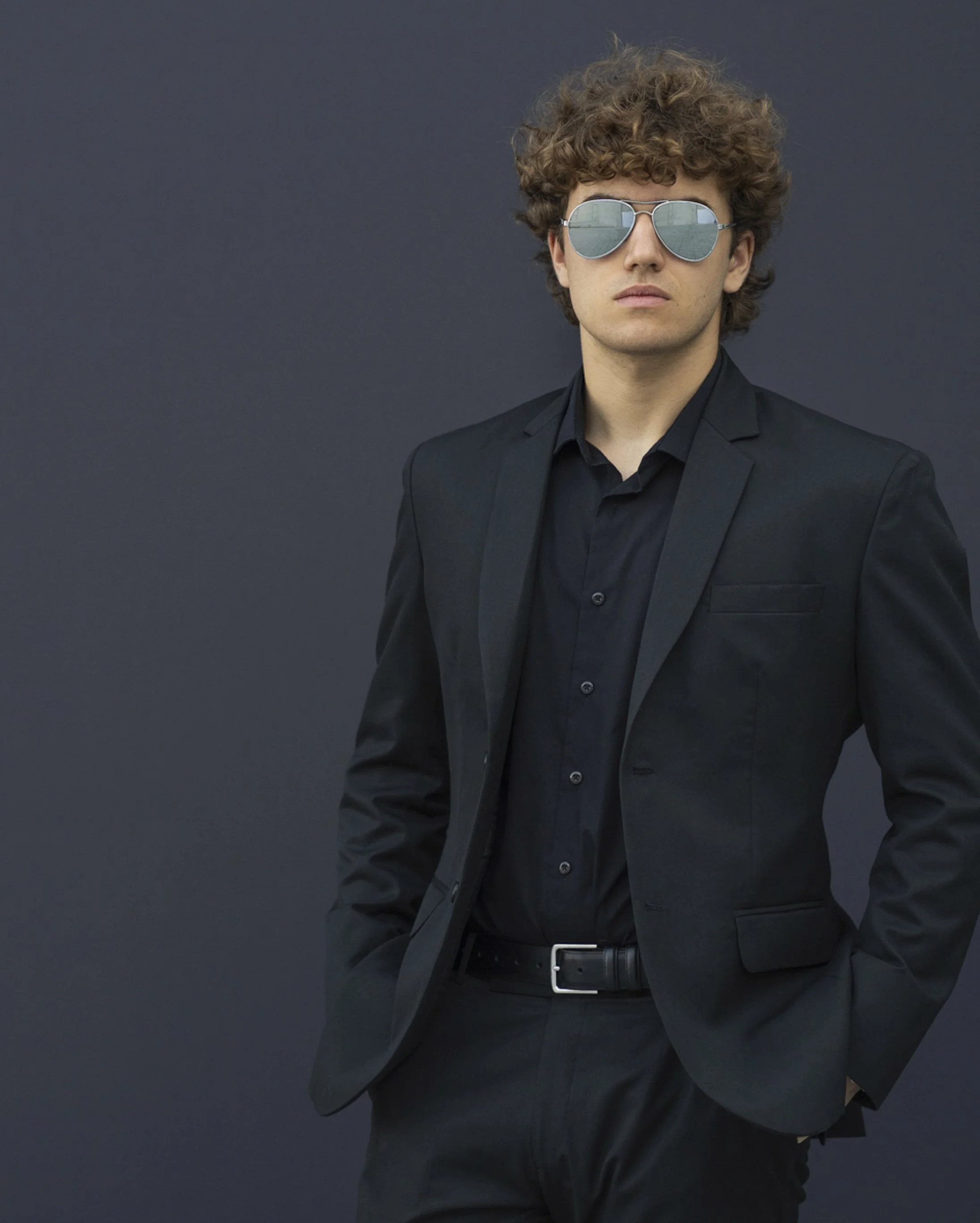A young man with curly hair and sunglasses dressed in a black suit, posing against a dark background.
