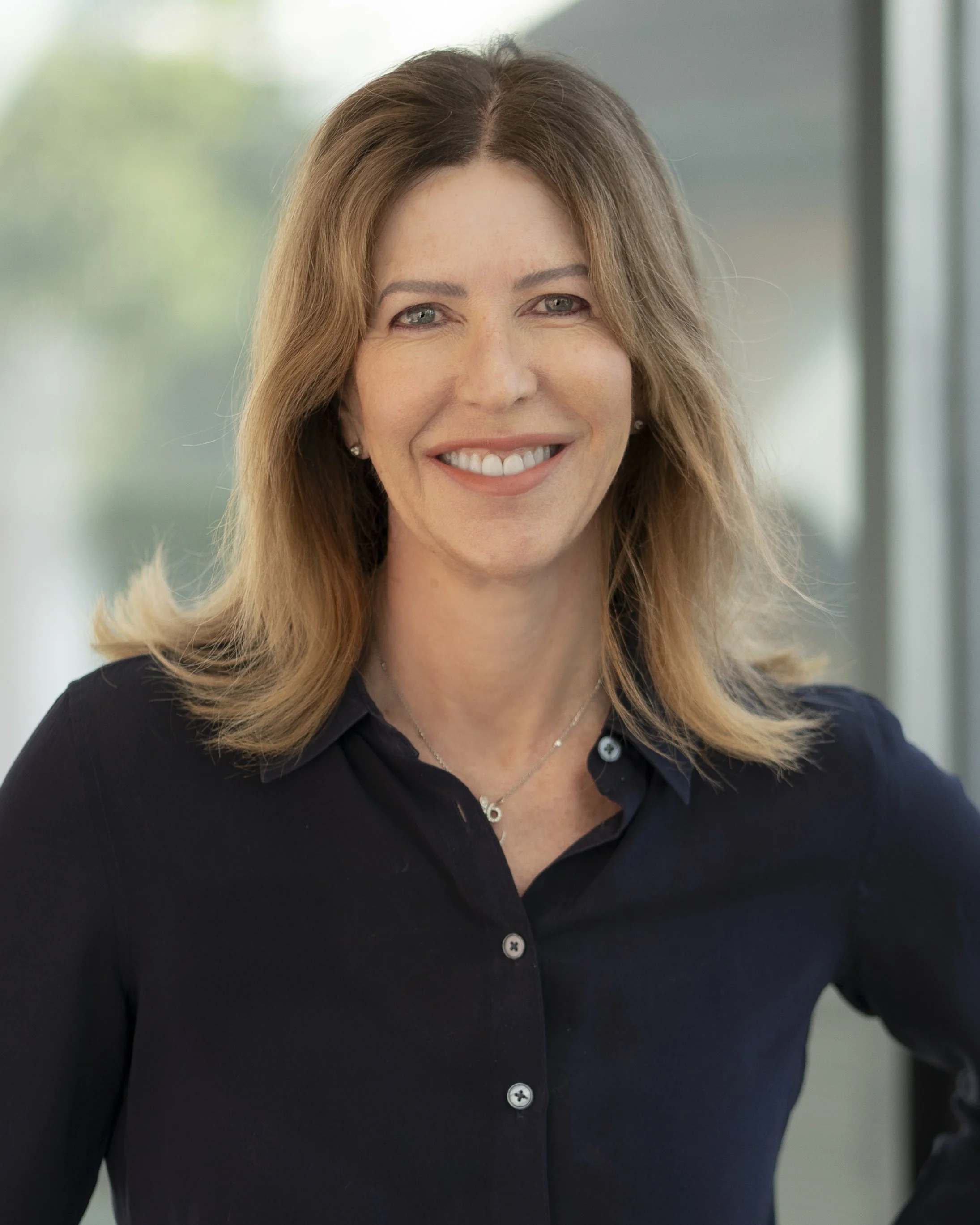 A woman with shoulder-length light brown hair, smiling, wearing a black collared shirt and a delicate necklace, standing indoors with a blurred background of windows and greenery.