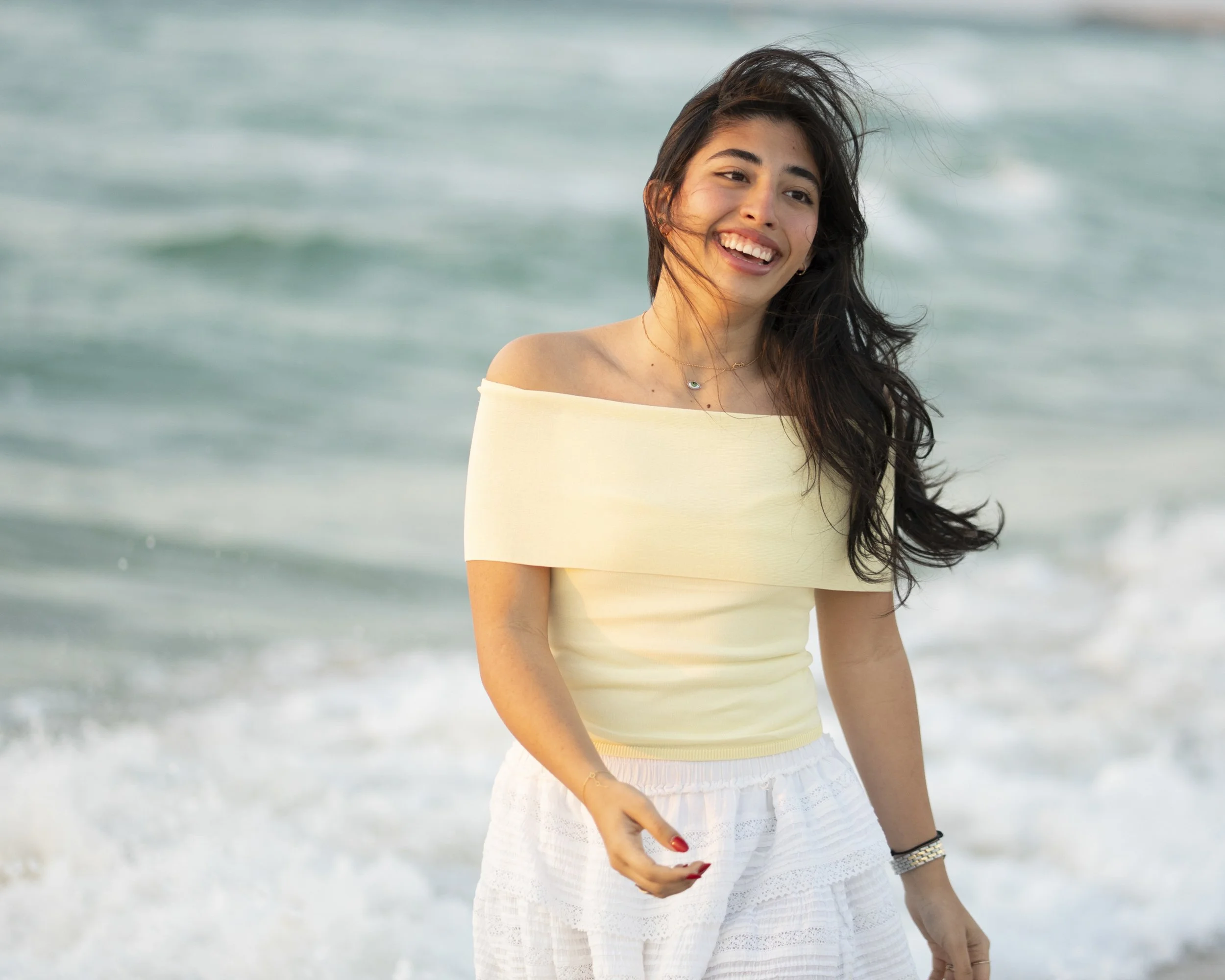 A smiling woman with long dark hair standing on the beach in front of the ocean, wearing a light yellow off-shoulder top and white skirt.