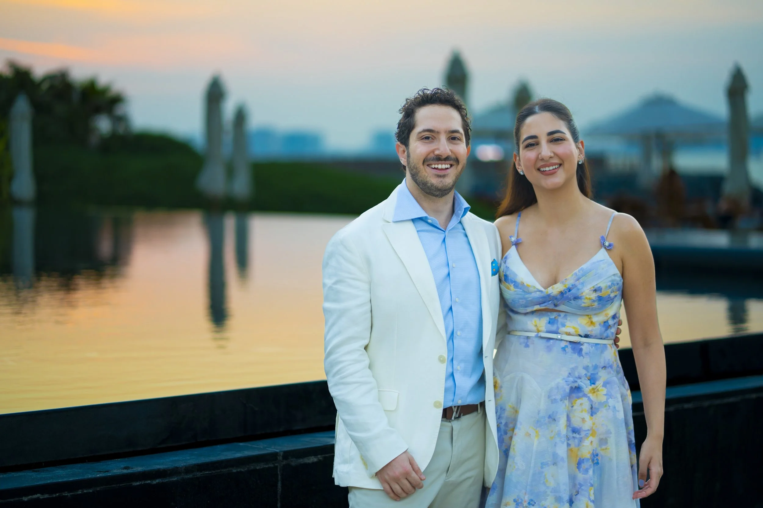 A smiling man and woman posing by a waterfront during sunset, with the man wearing a white blazer over a blue shirt and the woman in a pastel floral dress.