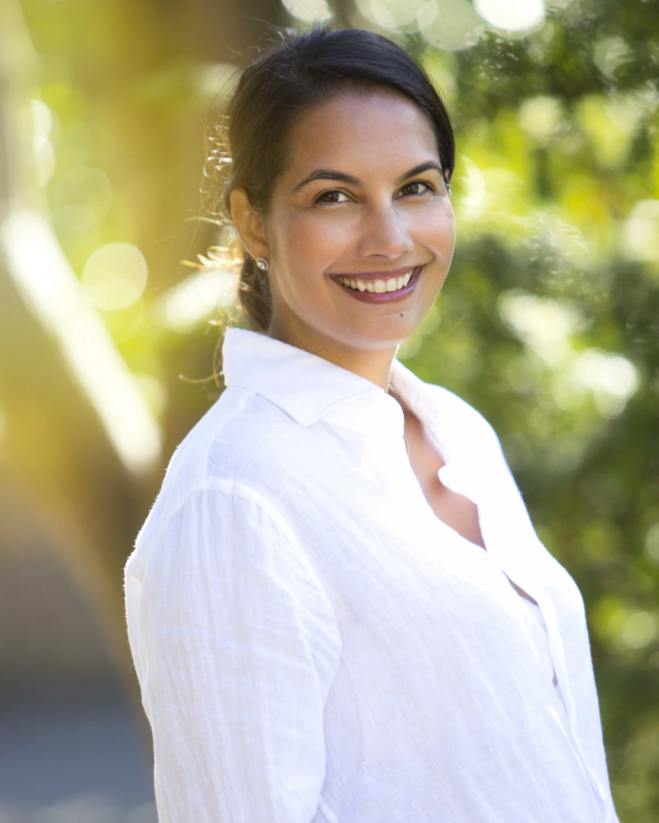 A woman with dark hair smiling in a white shirt outdoors with sunlight filtering through green trees.