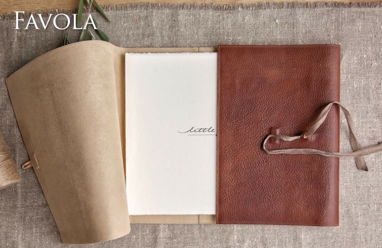 Open leather-bound notebook with blank pages and a red textured leather sleeve. The notebook is on a textured beige fabric surface, with a small green plant in the top left corner.