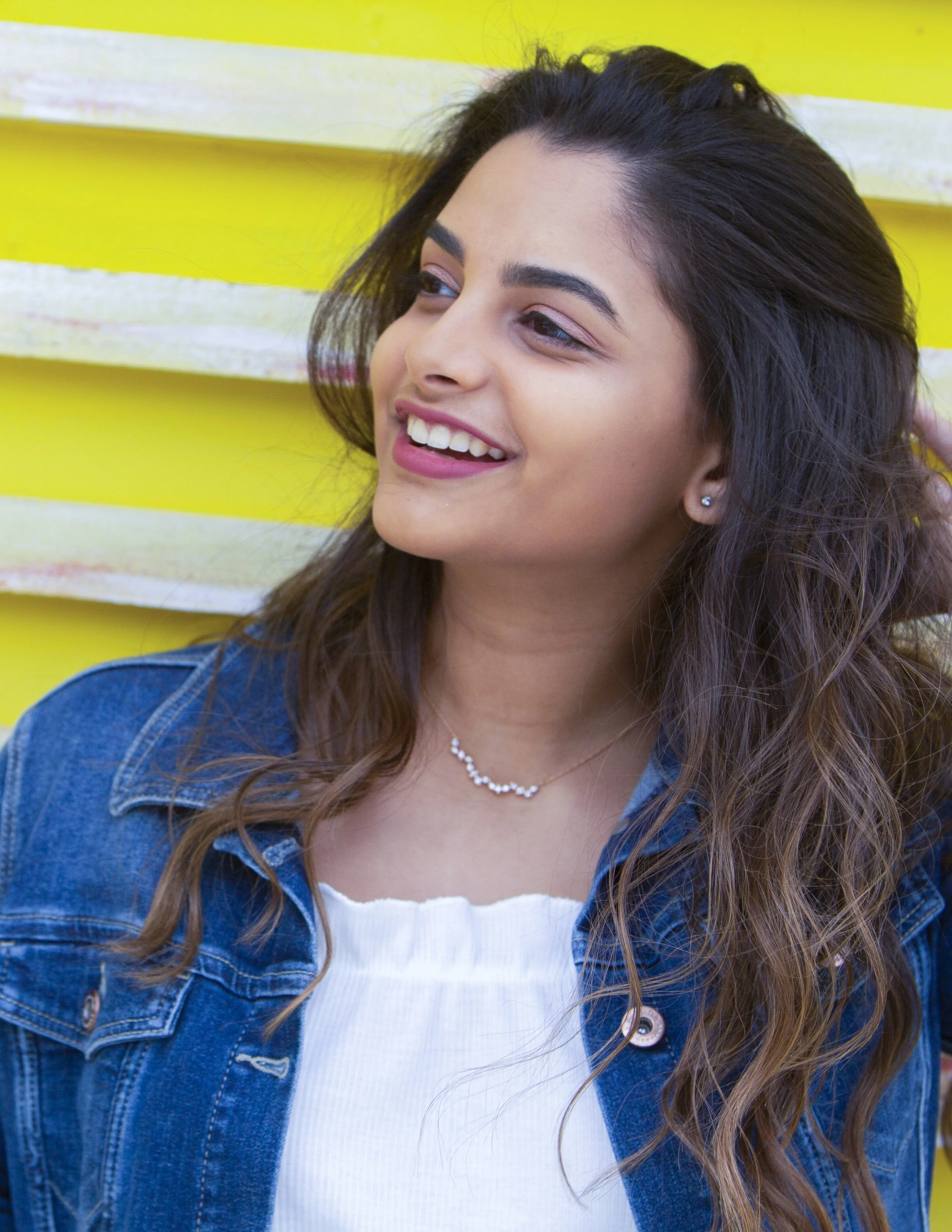 A young woman with long, wavy brown hair smiling and looking to her right, wearing a denim jacket, white top, and pearl necklace, standing against a bright yellow background.