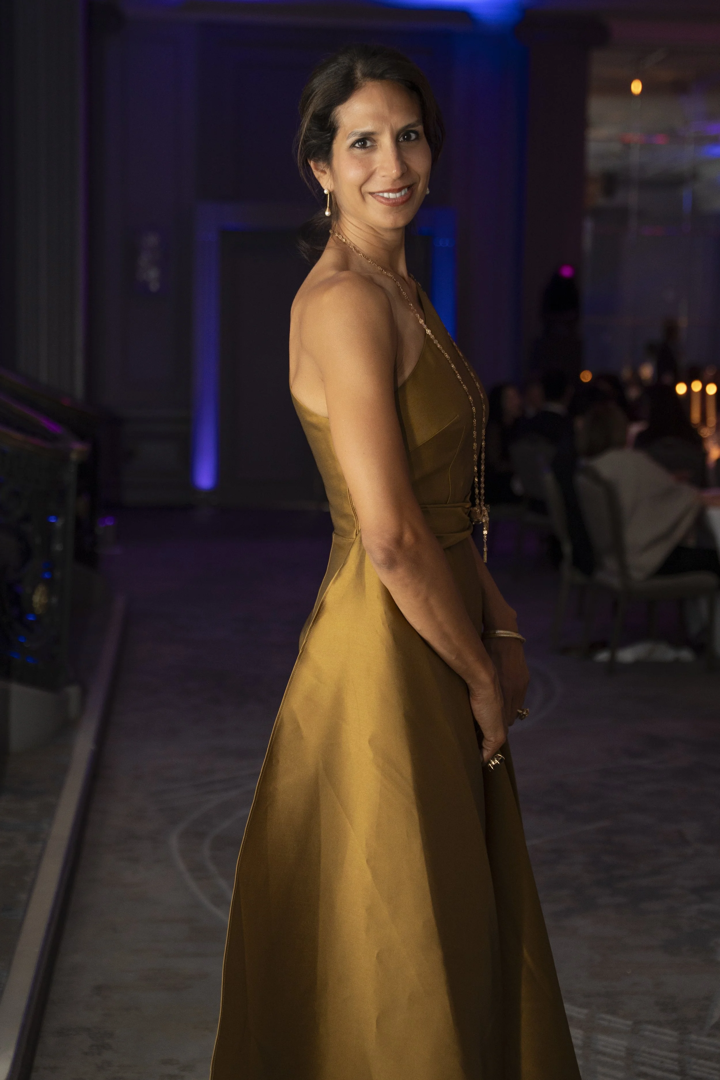 A woman in an elegant gold dress standing at a formal event, smiling at the camera with seated guests and dim lighting in the background.