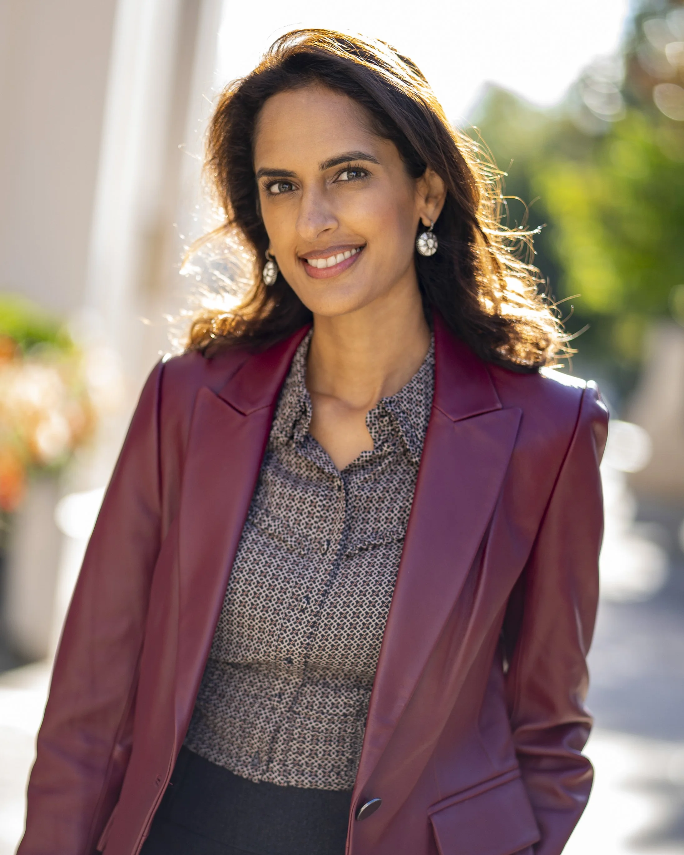 A woman with dark hair smiling outdoors wearing a maroon blazer, patterned blouse, and pearl earrings.