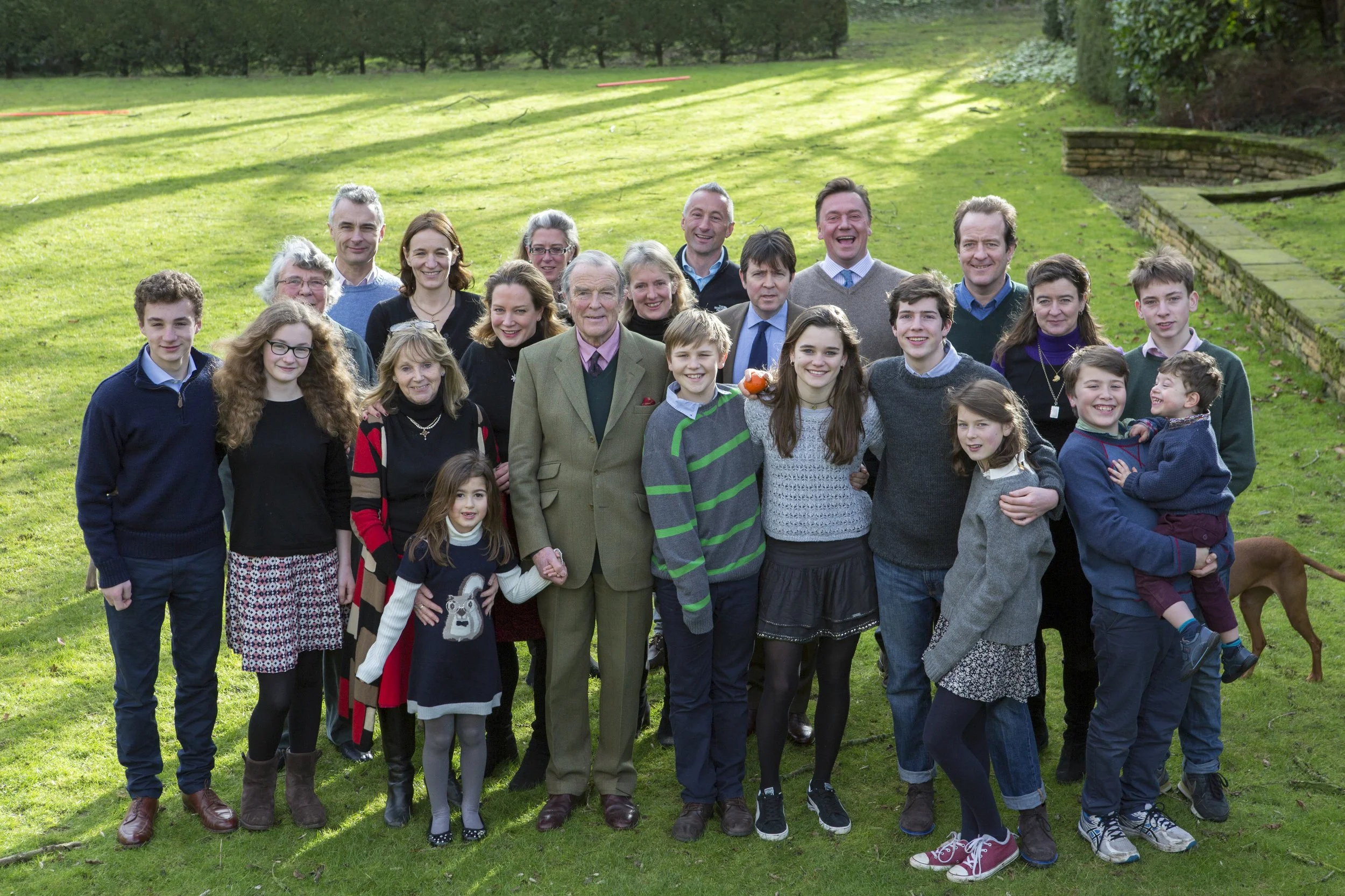 A large group of people, including children and adults, standing outdoors on a grassy area with trees in the background. They are smiling and posing for a group photo.