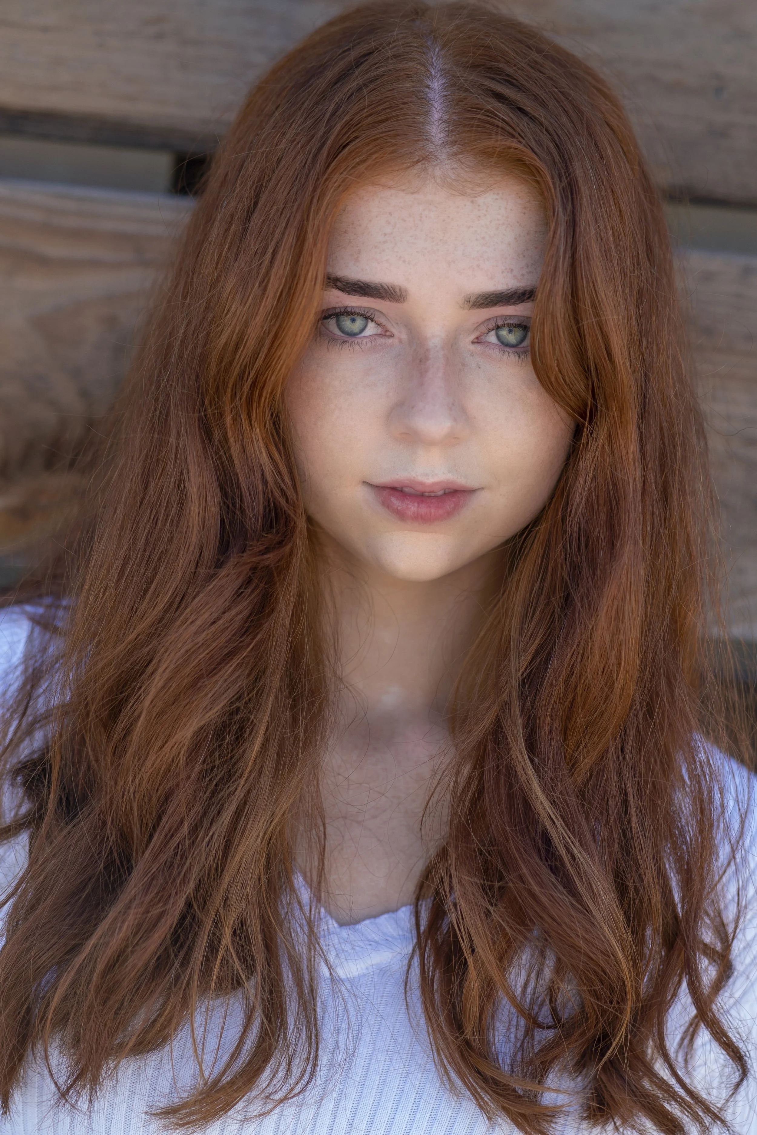 Close-up of a young woman with long, wavy red hair and blue eyes, wearing a white top, standing against a wooden background.