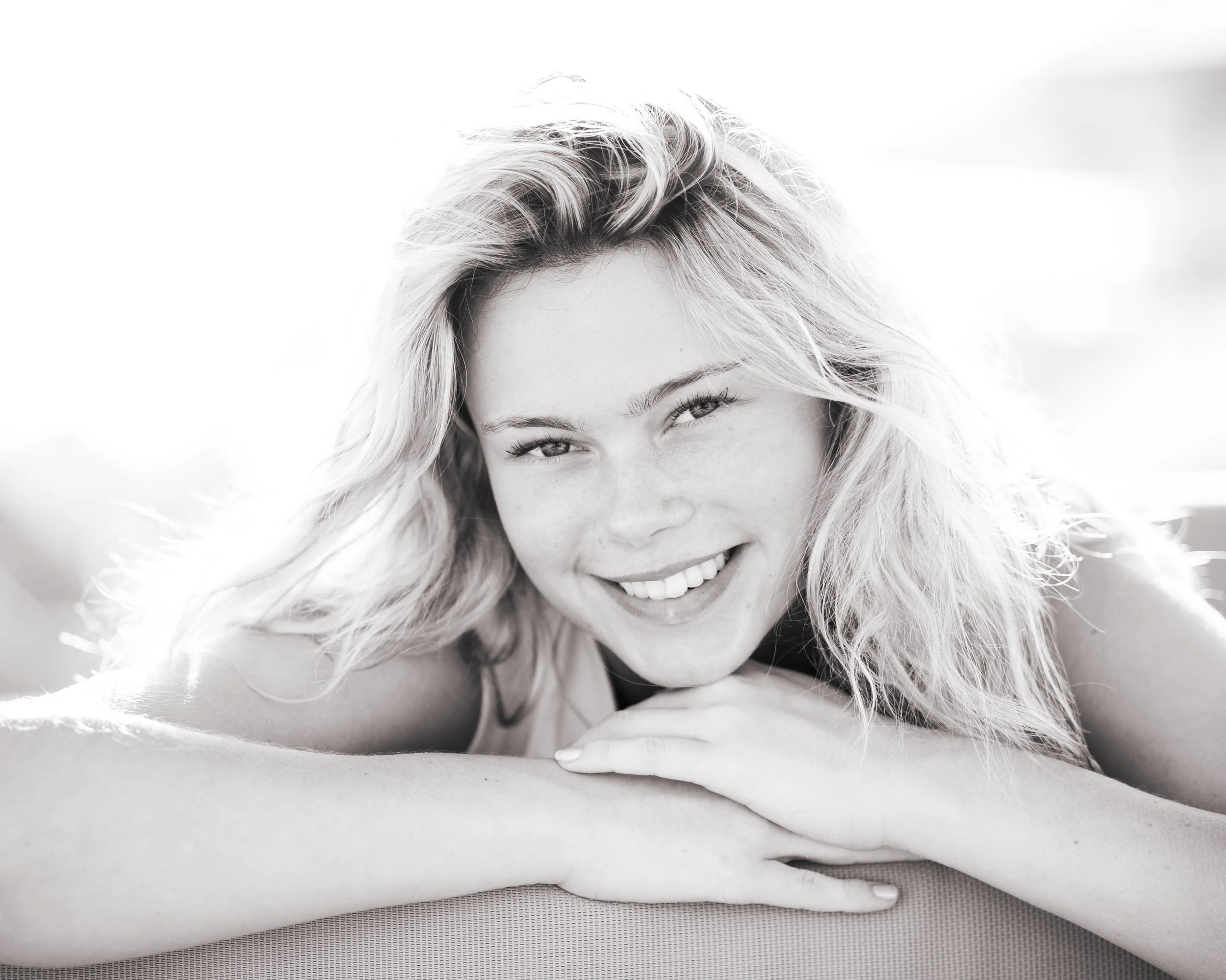 A woman with wavy hair lying on her stomach on a beach, smiling with her head resting on her arms.