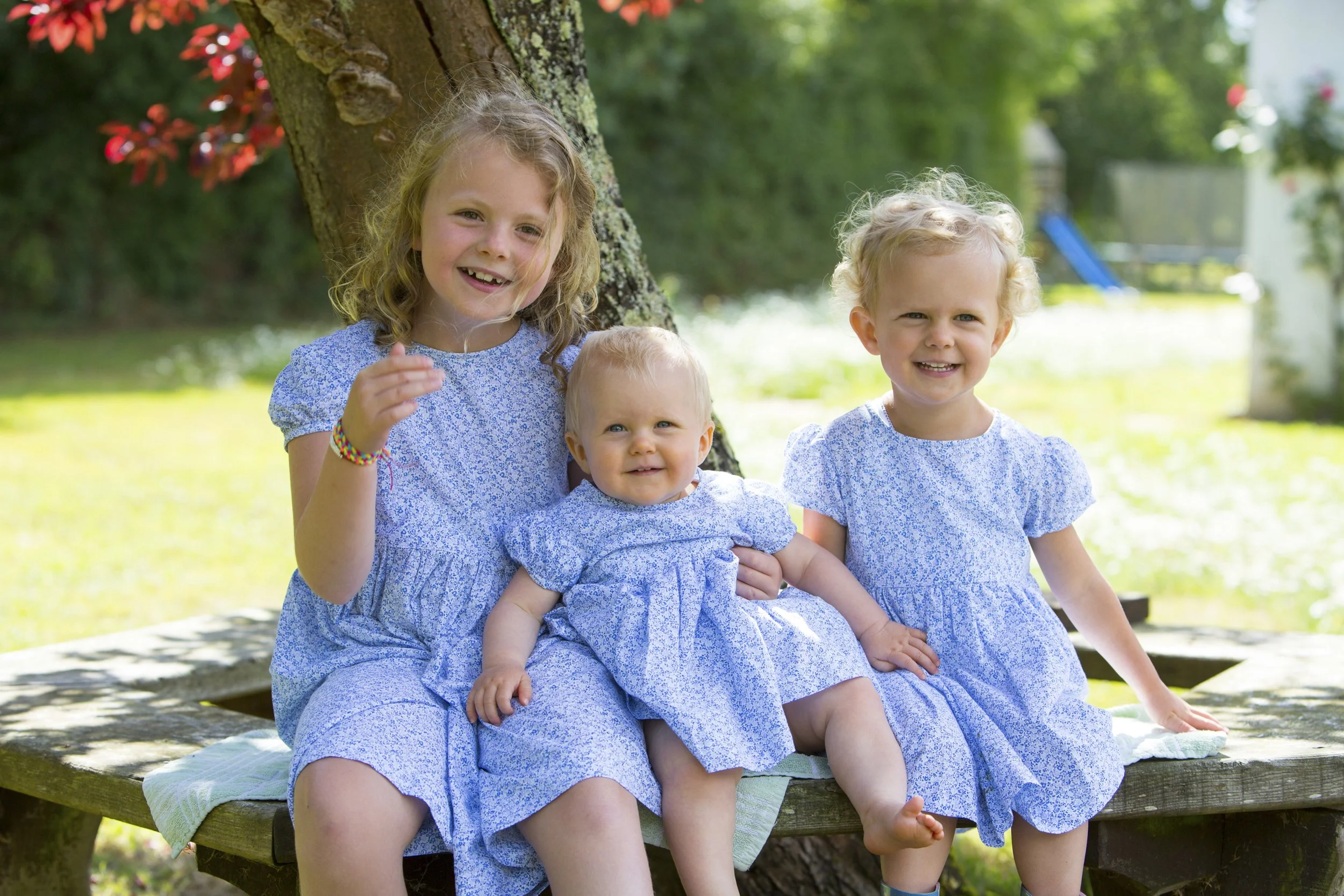 Three young girls sitting on a wooden bench outdoors, smiling, in a sunny garden with green grass and trees in the background.