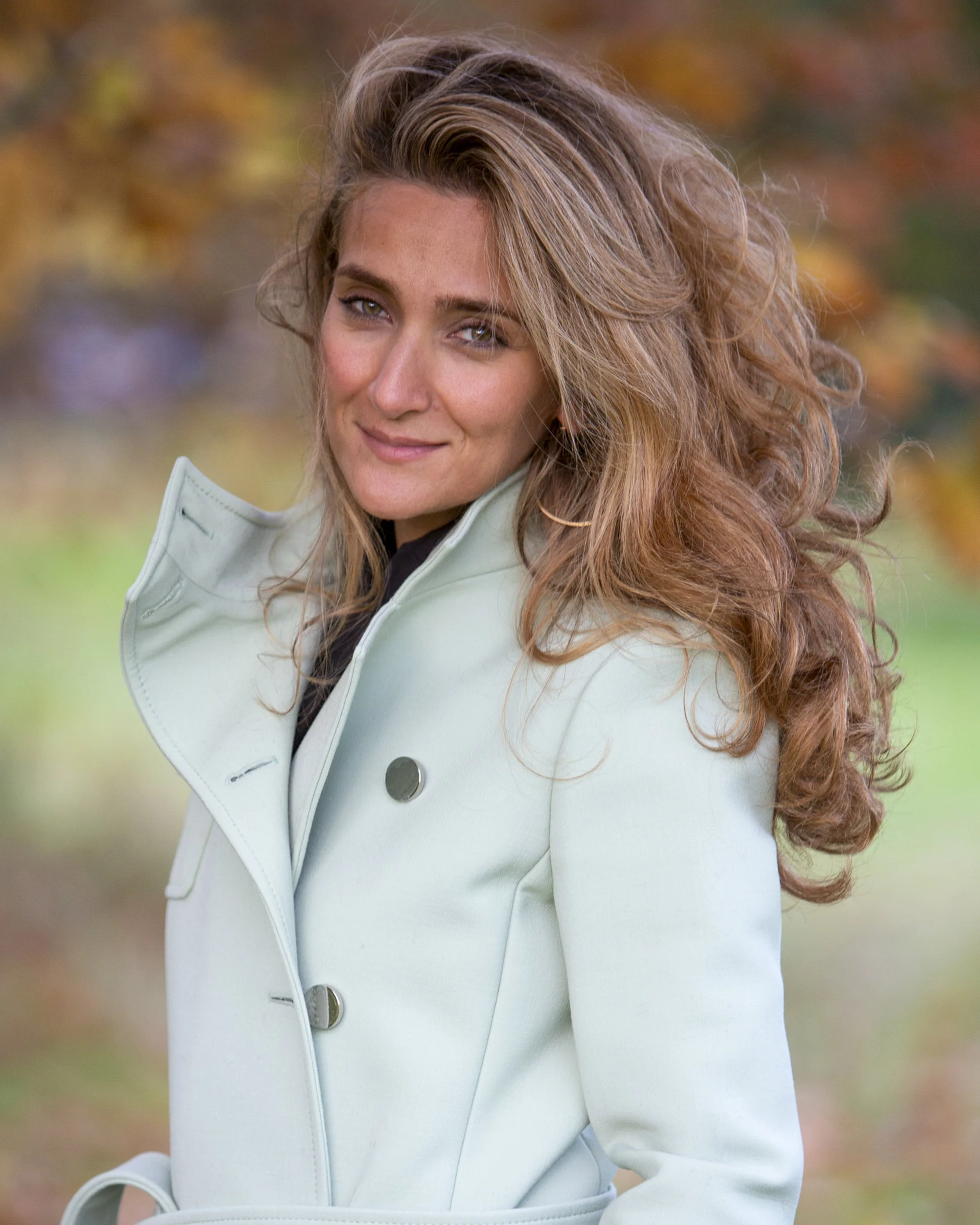 A woman with long, wavy light brown hair and blue eyes smiling outdoors in fall, wearing a light-colored coat.
