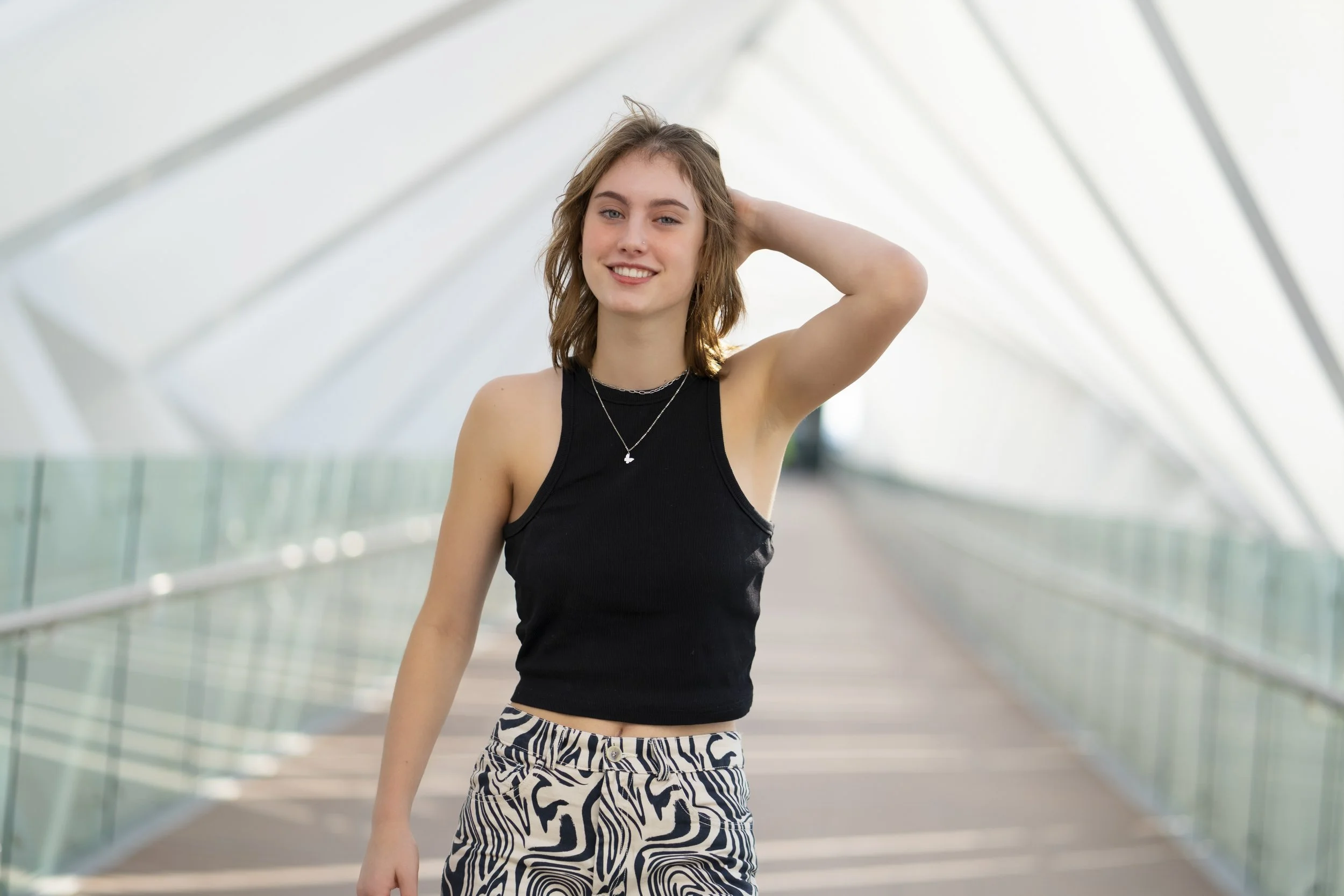 Young woman smiling in a modern glass walkway, wearing a black sleeveless top and patterned pants.