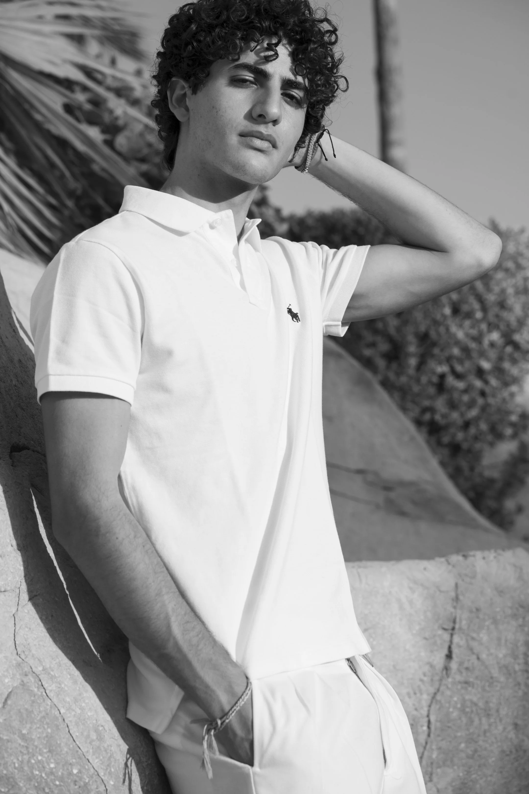 Black and white photo of a young man with curly hair, wearing a white polo shirt with a small logo, standing outdoors against rocks and trees, looking at the camera with a serious expression.