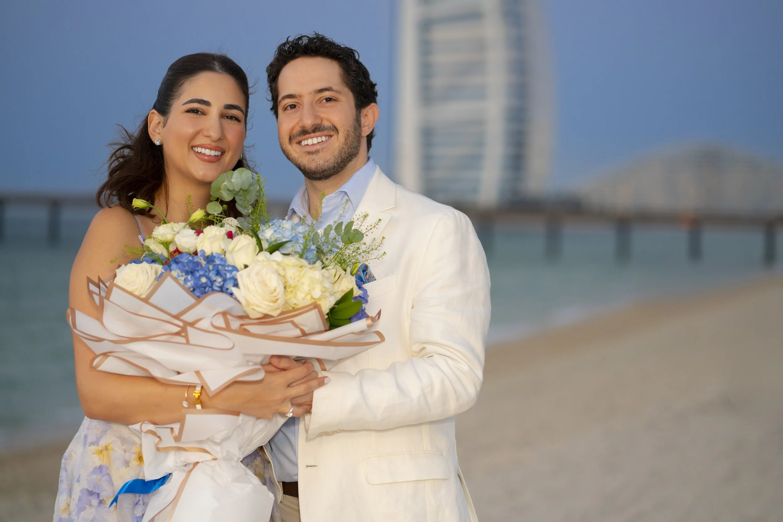 A happy couple on the beach, with a city skyline and pier in the background, holding a bouquet of flowers and smiling at the camera.