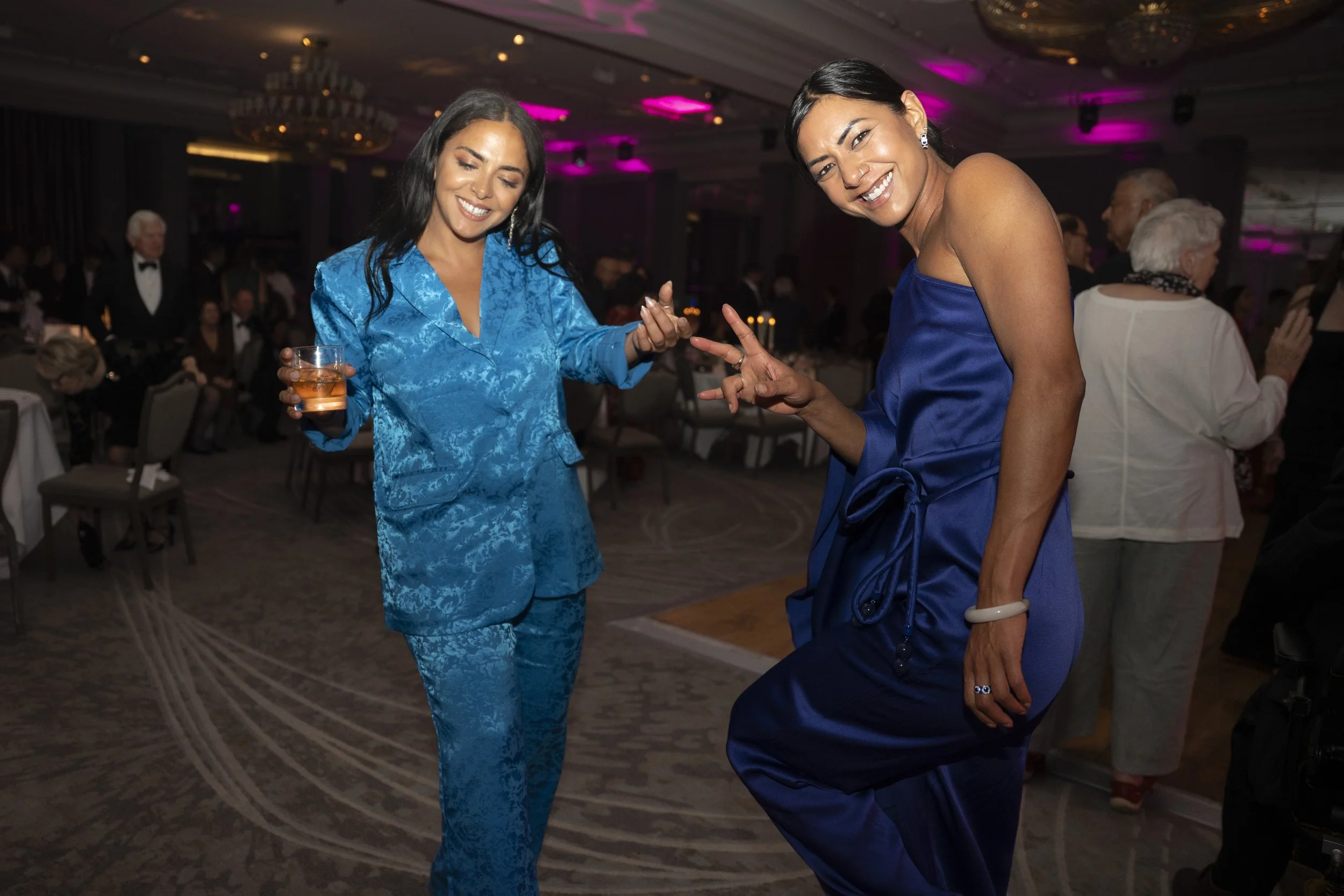 Two women dancing and smiling at an indoor event, with people seated at tables in the background. Both women are wearing blue outfits, one holding a drink and the other with a bracelet and earrings.