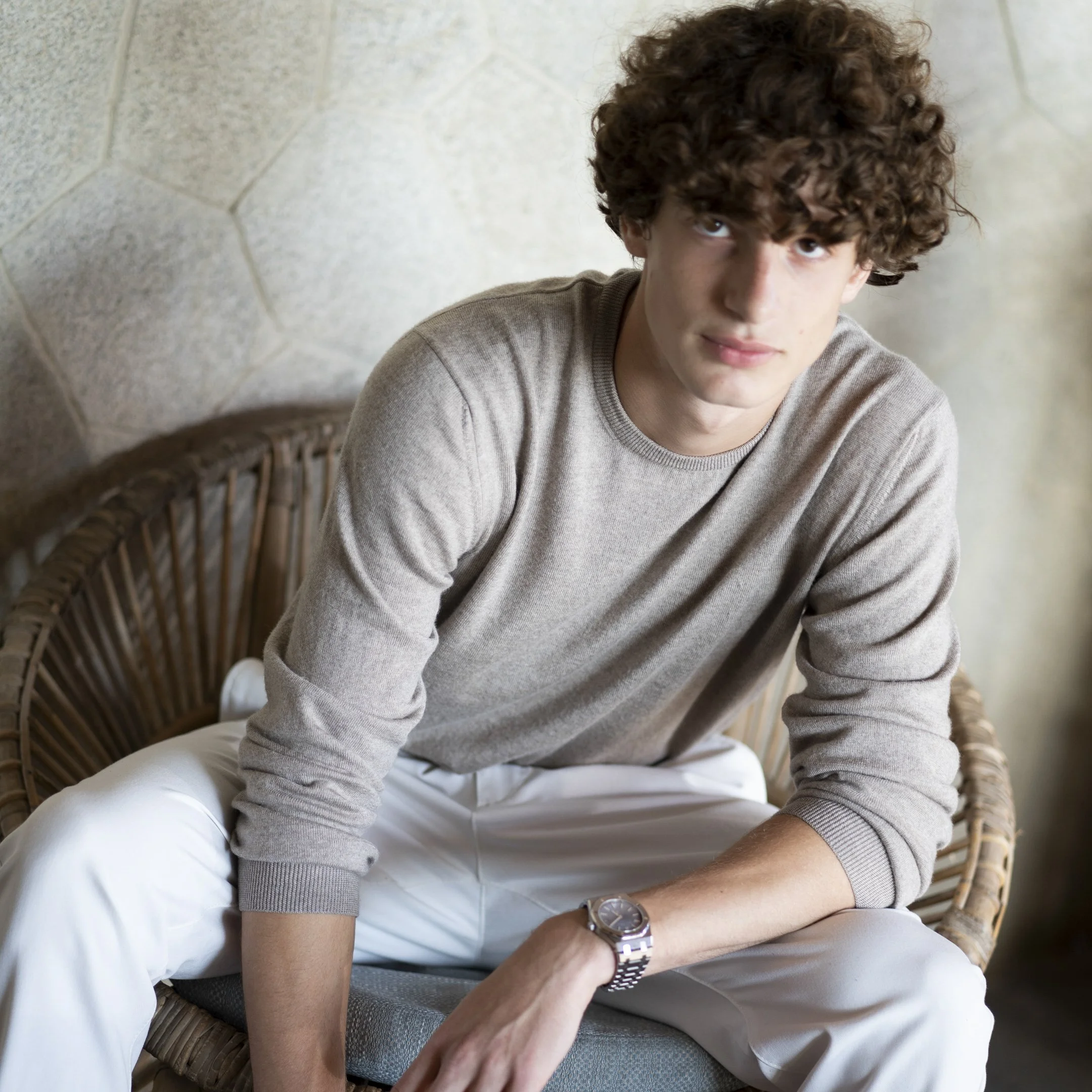 Young man with curly hair wearing a beige sweater and white pants, sitting on a wicker chair with a stone wall in the background.
