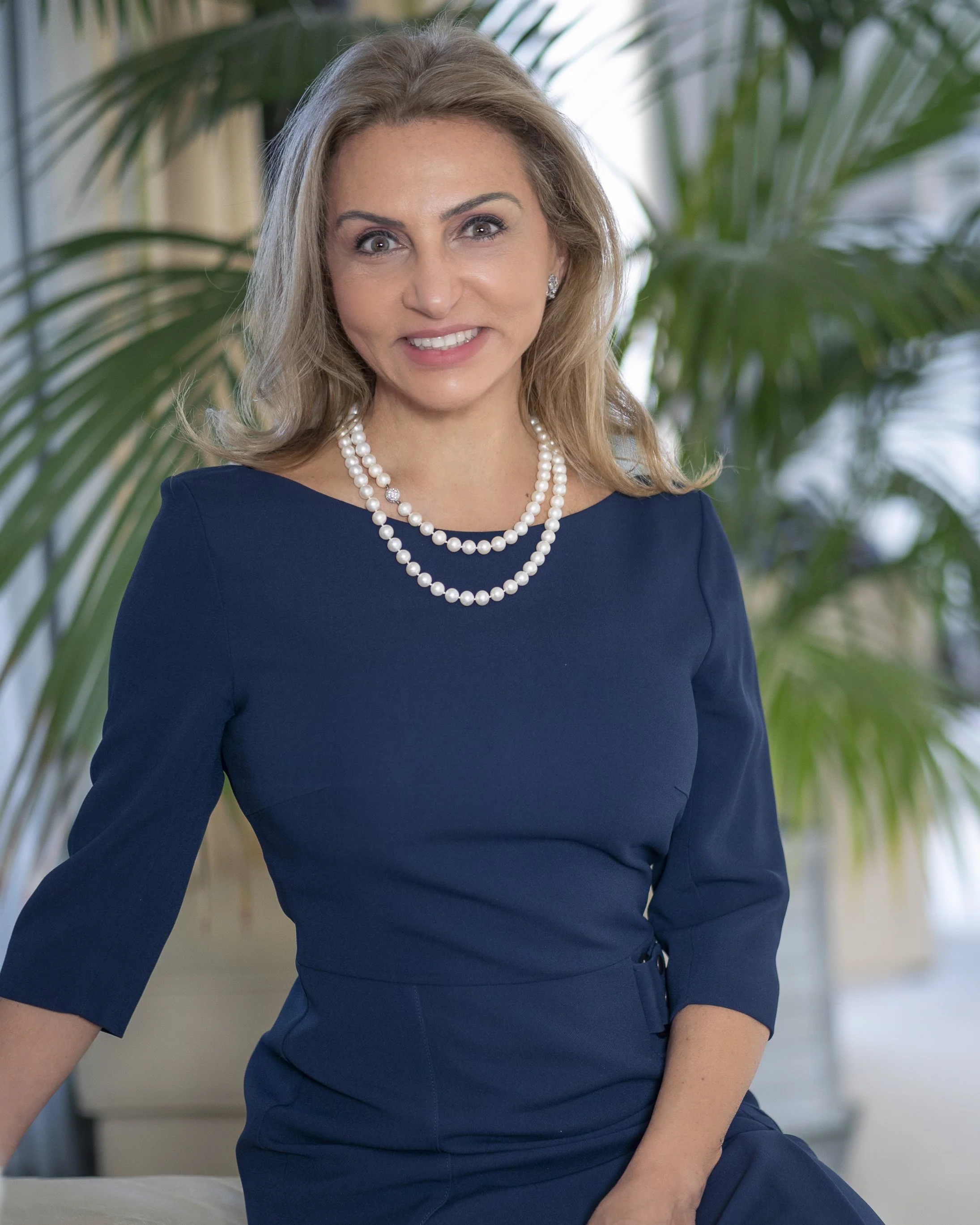 A woman in a navy blue dress with a double string of pearls around her neck, smiling, with a background of green plants.