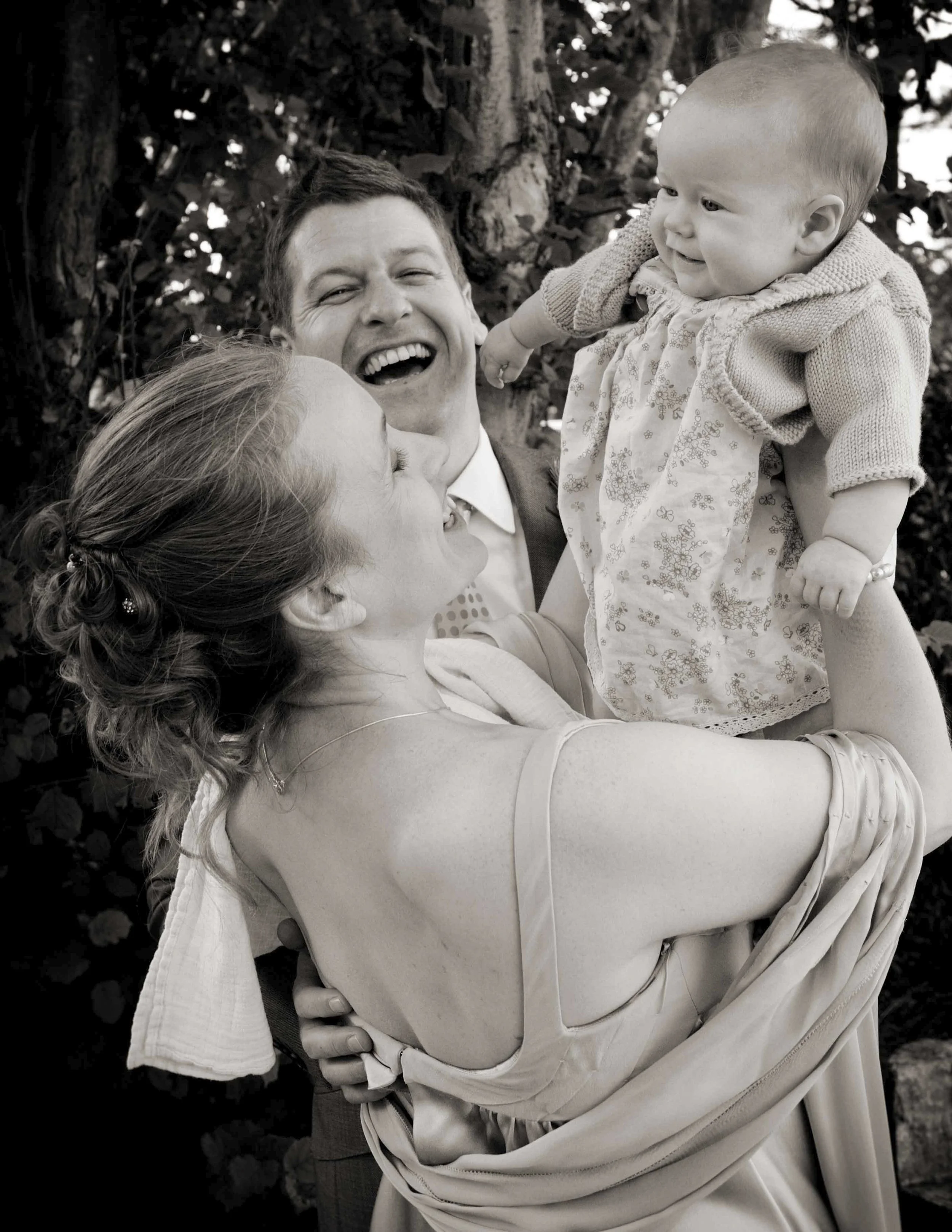 A smiling woman in a wedding dress being lifted by a man in a suit, holding a young child, with a tree in the background.