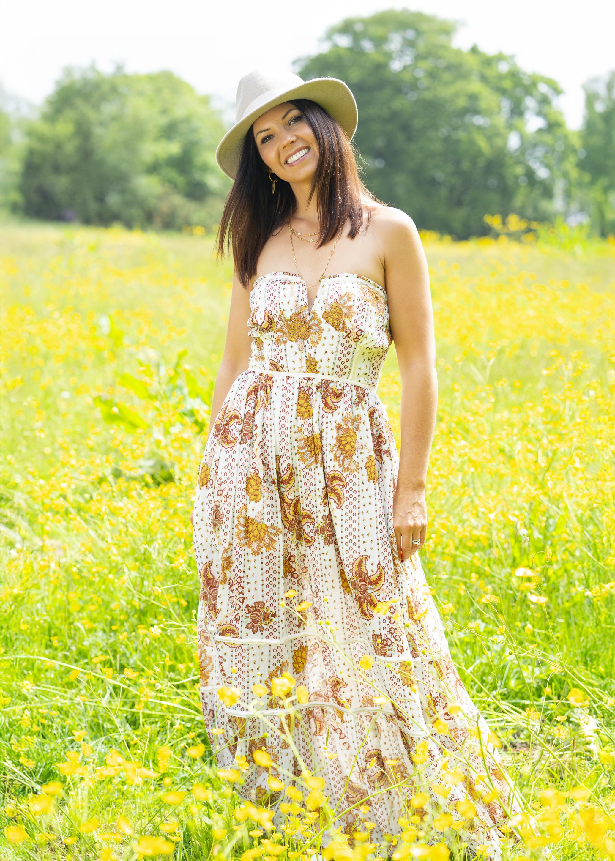 A woman in a strapless floral dress and wide-brimmed hat standing in a field of yellow flowers with green trees in the background.