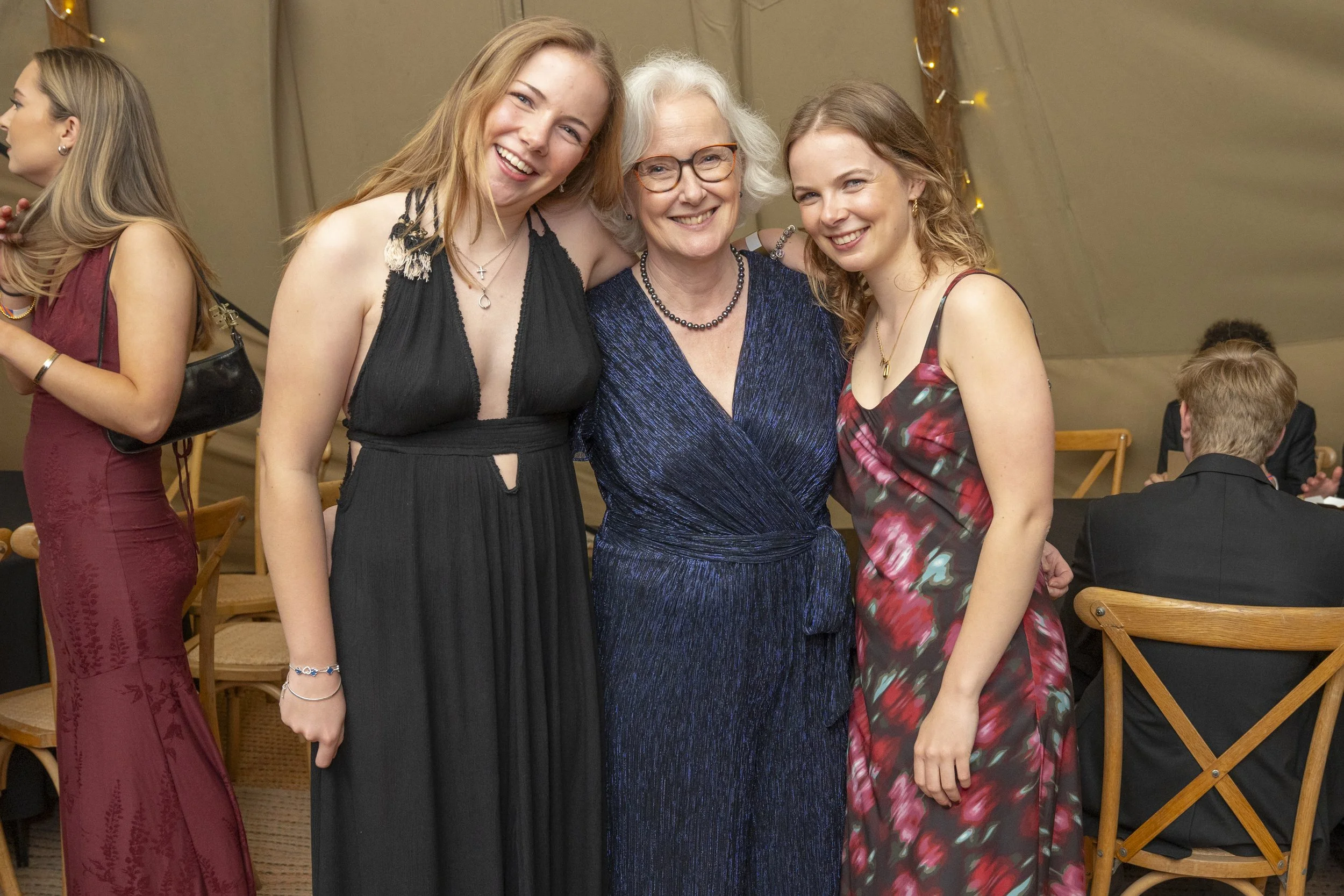 Three women smiling at a social event, dressed in evening wear, standing close together with arms around each other, with a woman in a black dress on the left, an older woman in a blue dress in the middle, and a woman in a floral dress on the right.