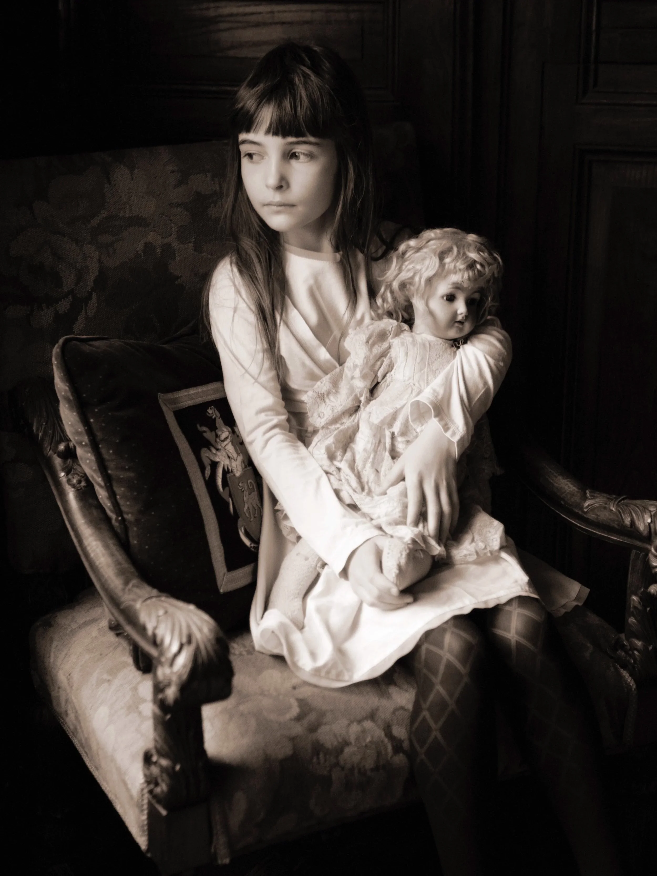 A girl with long dark hair and bangs sitting on an ornate vintage armchair, holding a porcelain doll dressed in vintage clothing, in a dimly lit room with dark wood paneling.
