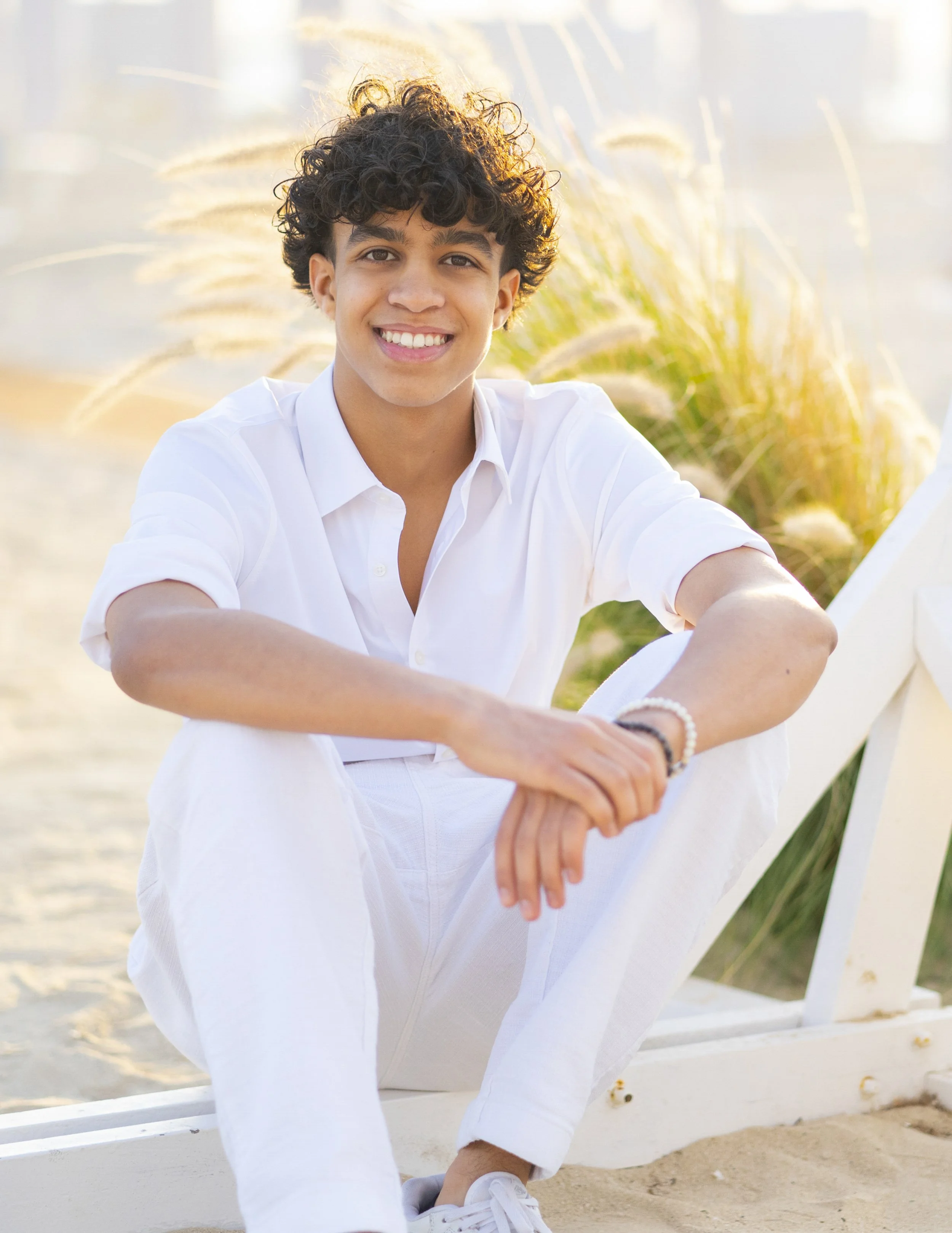 A young man with curly dark hair and a white shirt sitting on a white bench at the beach, smiling at the camera, with tall grasses in the background and soft sunlight.