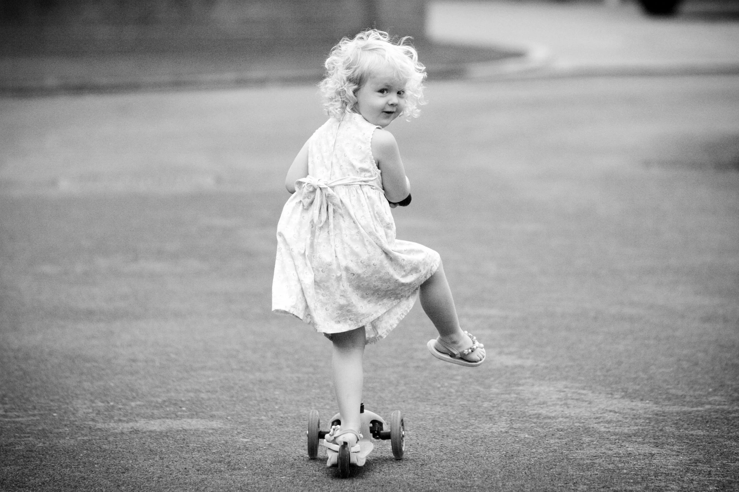 A young girl with curly hair riding a skateboard on a paved street, looking back over her shoulder.