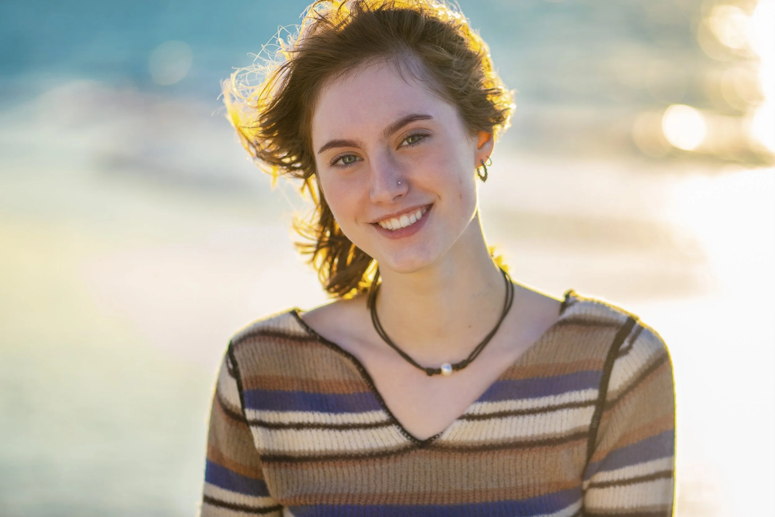A young woman with short, wavy brown hair smiling outdoors near water during sunset.