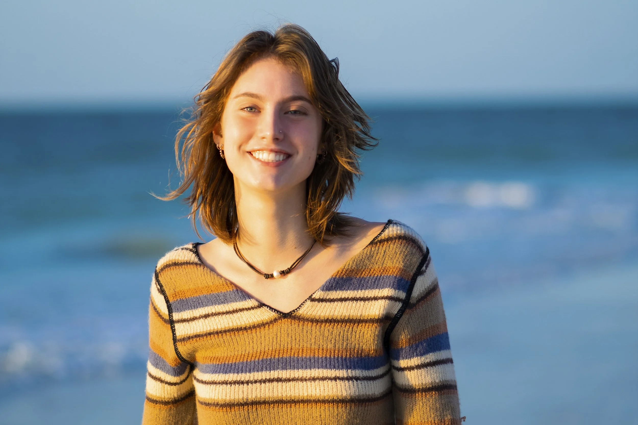 A young woman with shoulder-length brown hair smiling at the camera, standing outdoors near the ocean at sunset, wearing a striped sweater and necklace.