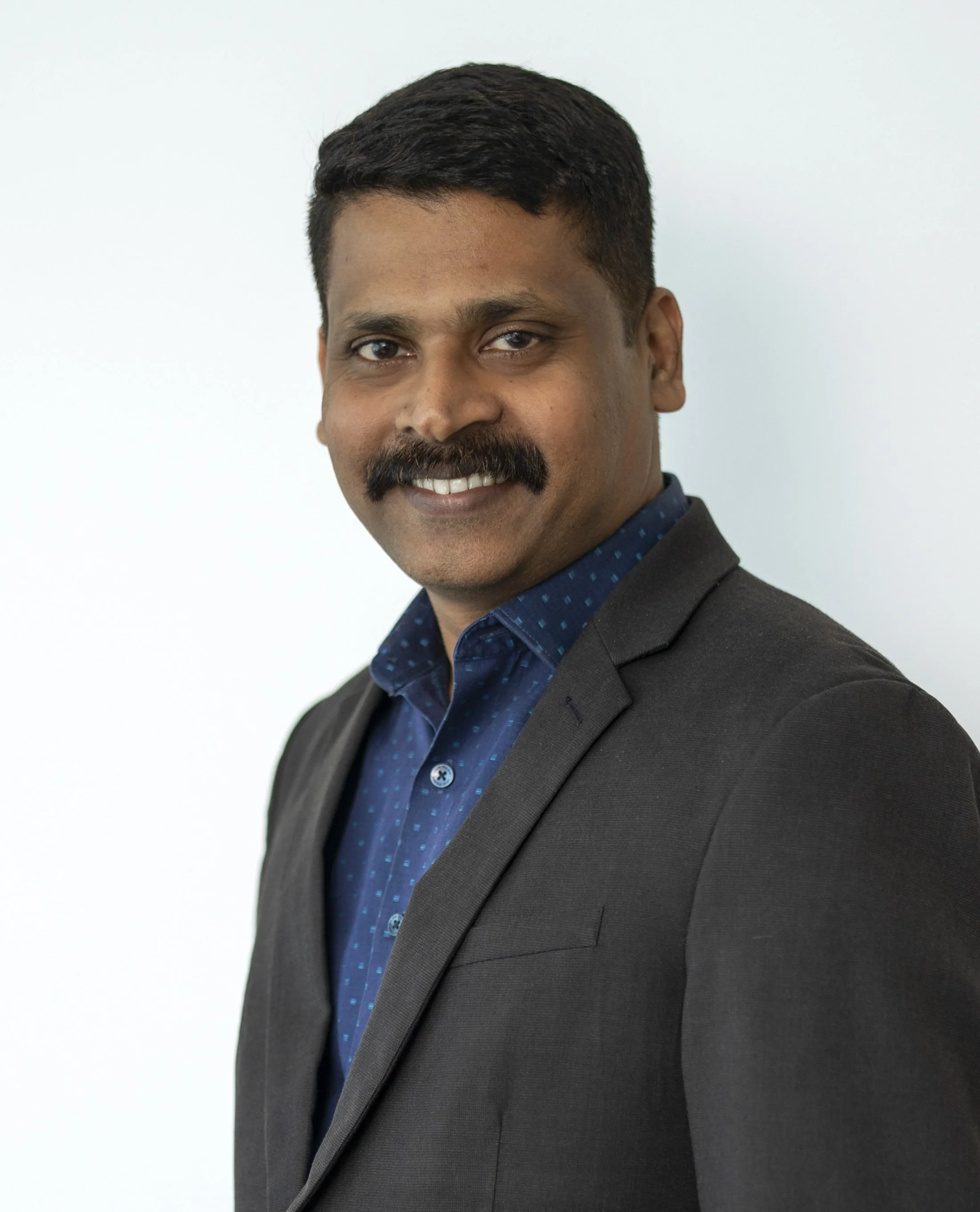 Professional portrait of a man with a mustache and short hair, wearing a dark suit jacket and a blue shirt with small dots, smiling against a plain white background.