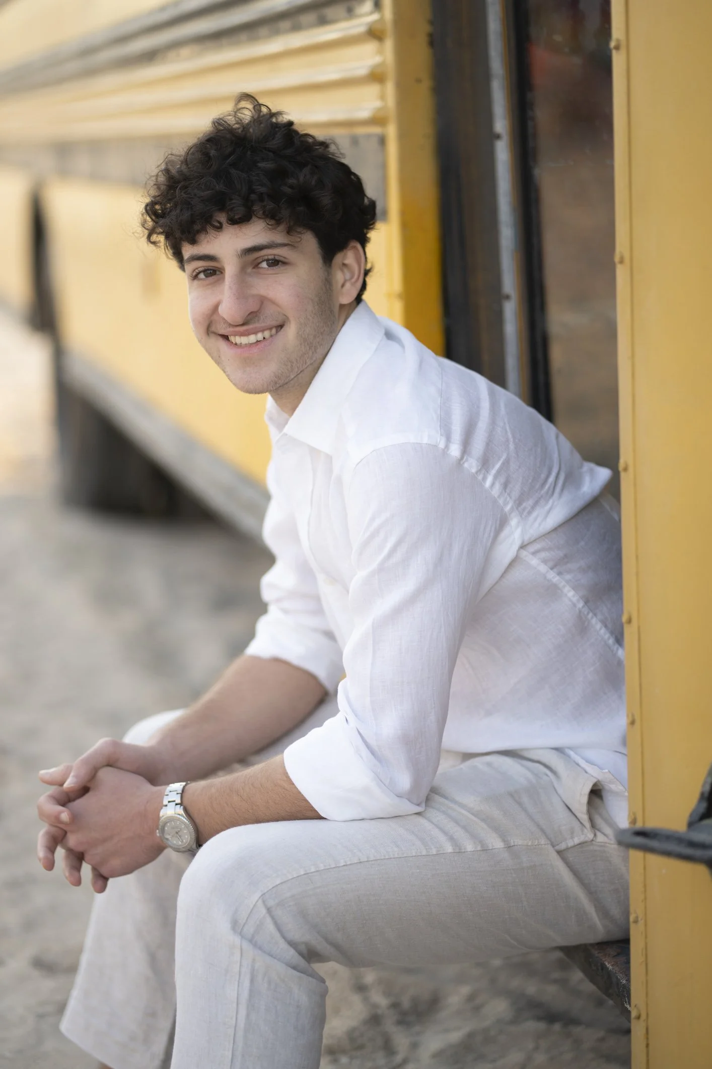 Young man with curly hair and a white shirt sitting on a bench outside a yellow train car, smiling at the camera.