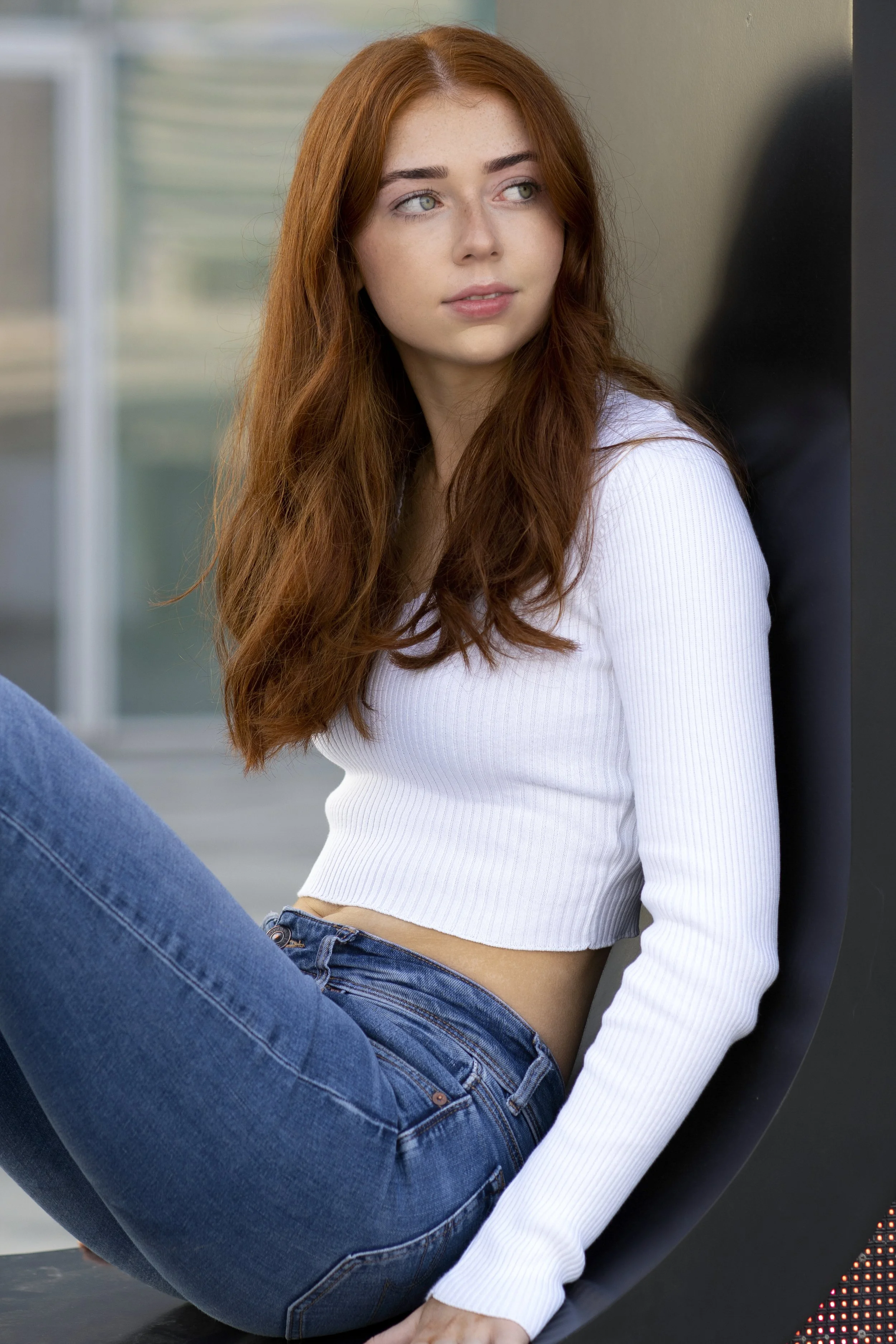 Young woman with long red hair, wearing a white cropped sweater and blue jeans, sitting against a black chair, looking away thoughtfully.