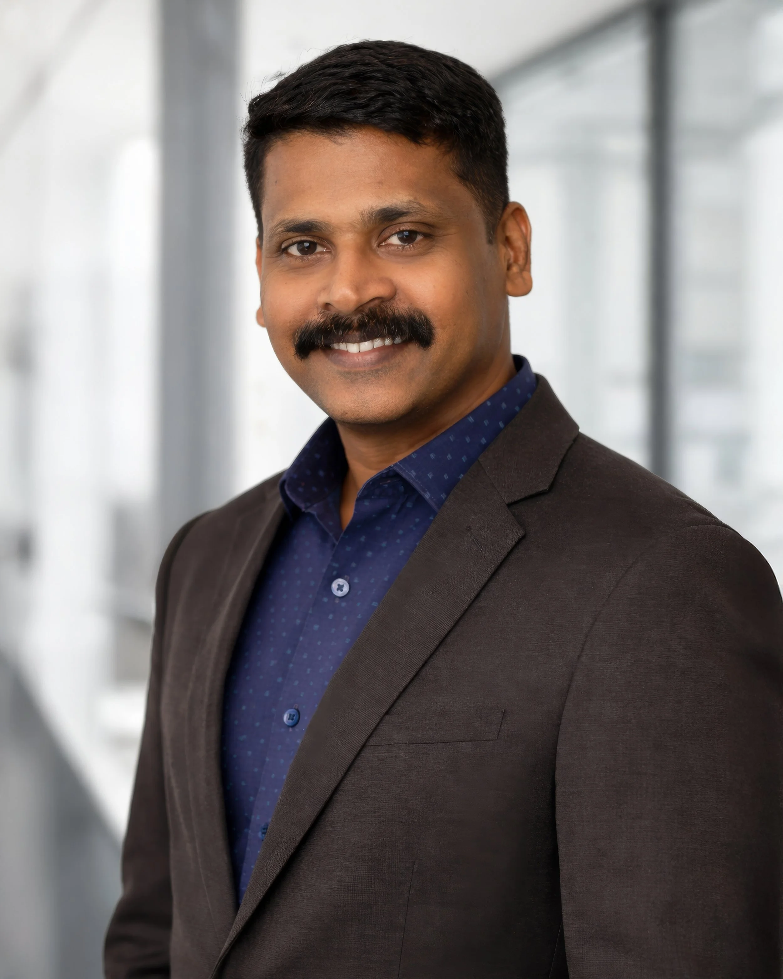 Professional headshot of a man with dark hair, mustache, wearing a dark blazer and blue shirt, smiling in an office setting with large windows.