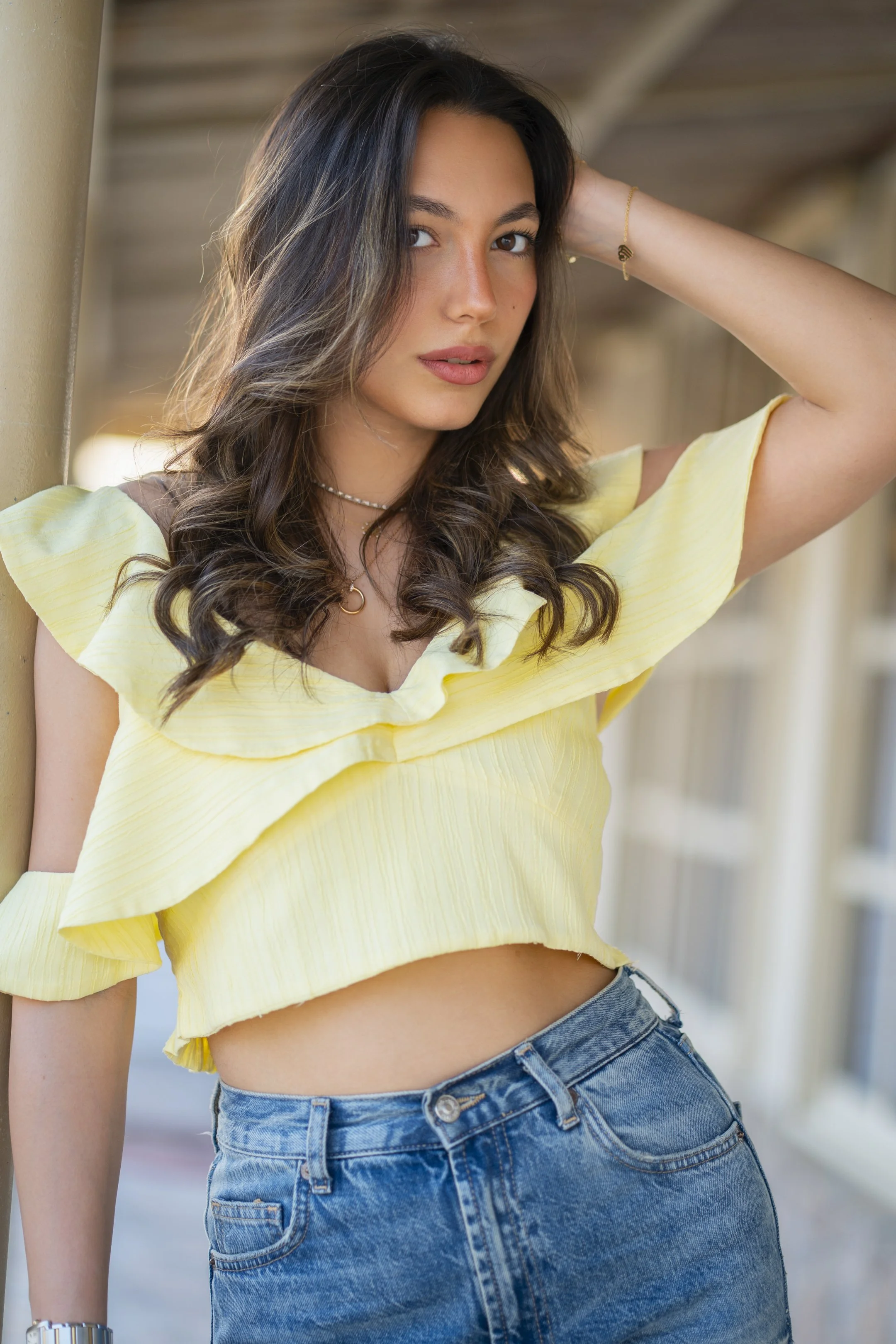 A young woman with long, wavy brown hair wearing a yellow ruffled crop top and high-waisted jeans, posing outdoors with her hand behind her head.