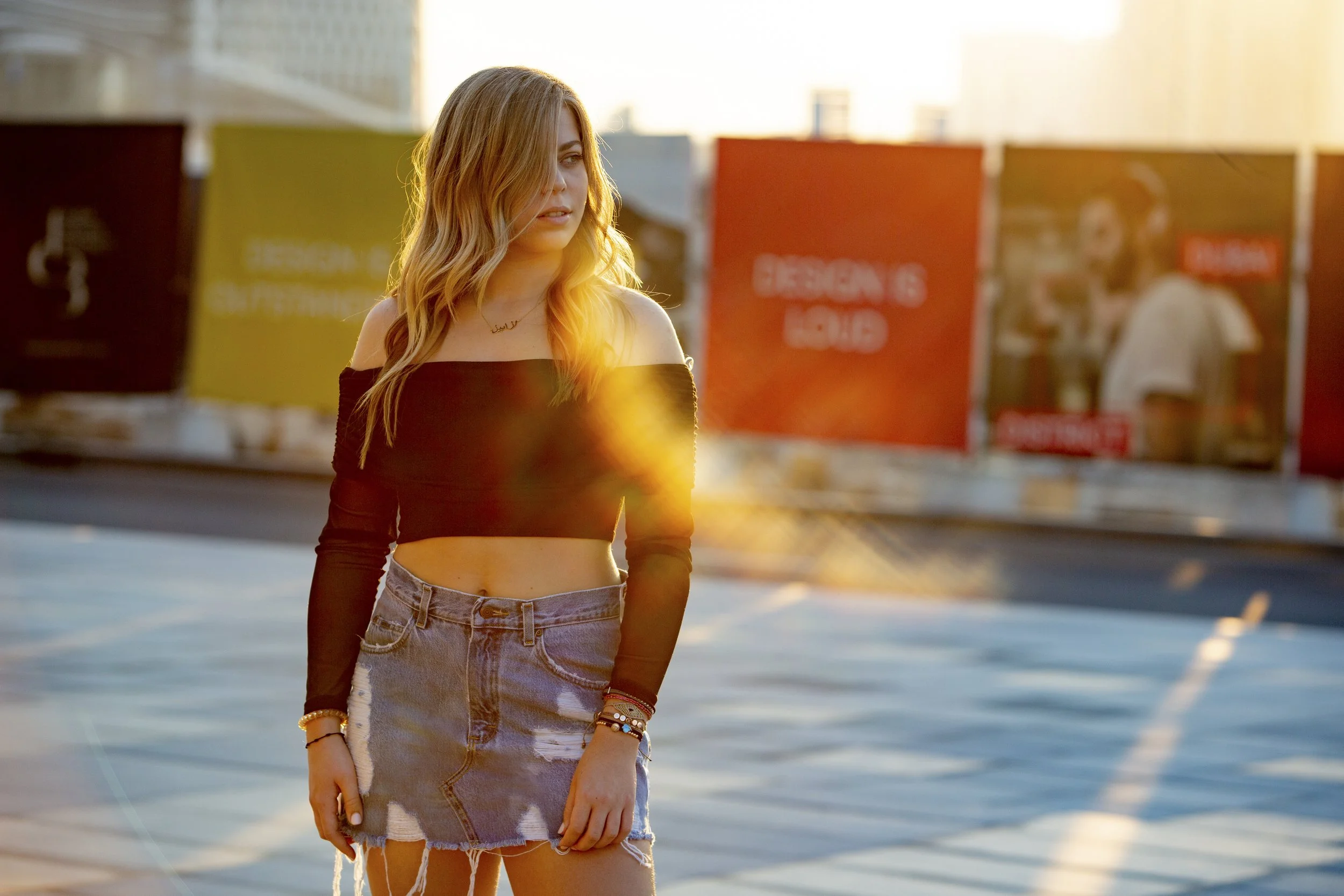 A young woman with long blonde hair wearing an off-the-shoulder black crop top and a denim skirt, standing on a rooftop parking lot with city buildings and billboards in the background during sunset.